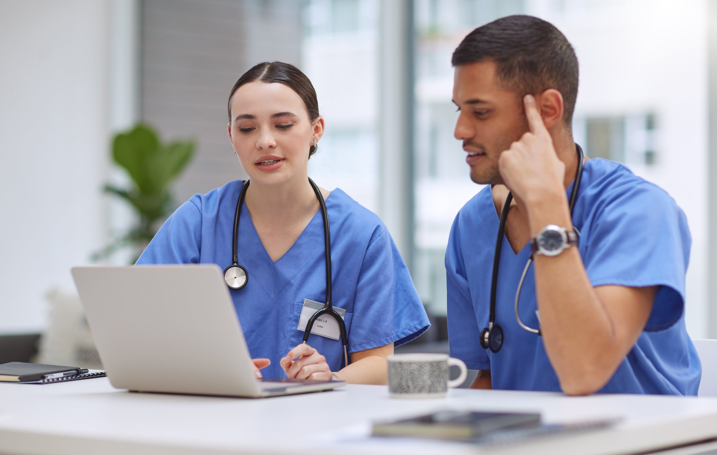 Two young nurses in blue scrubs sitting at a table and working on a computer together.