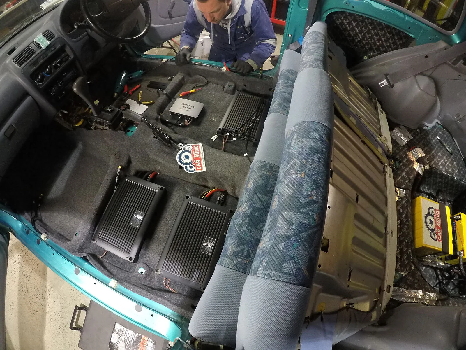 Person installing audio equipment inside a car, with seats removed and various components visible, including amplifiers and wiring.