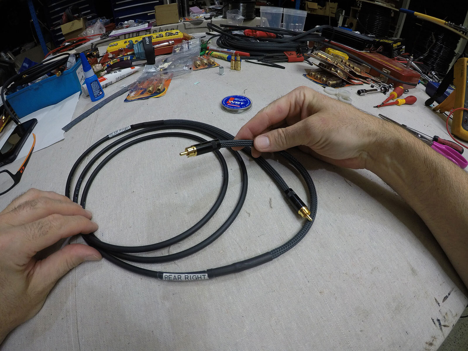 Hands holding coaxial cable labeled 'Rear Right' on a cluttered workbench with tools and electronic components visible.