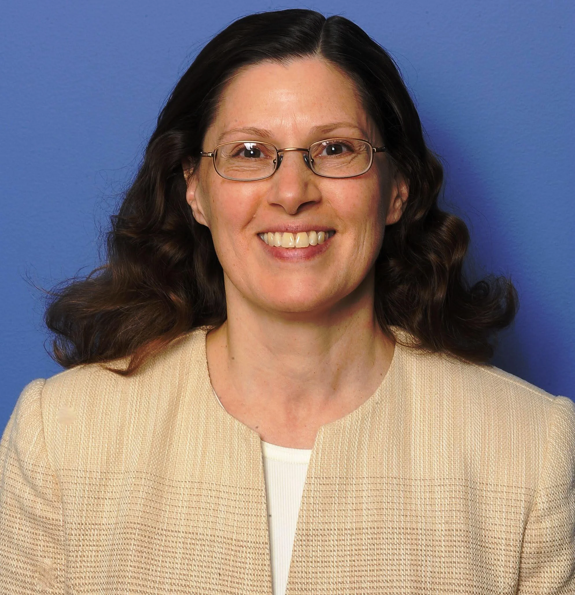 Close-up of a woman with glasses smiling, wearing a beige sweater against a blue background.