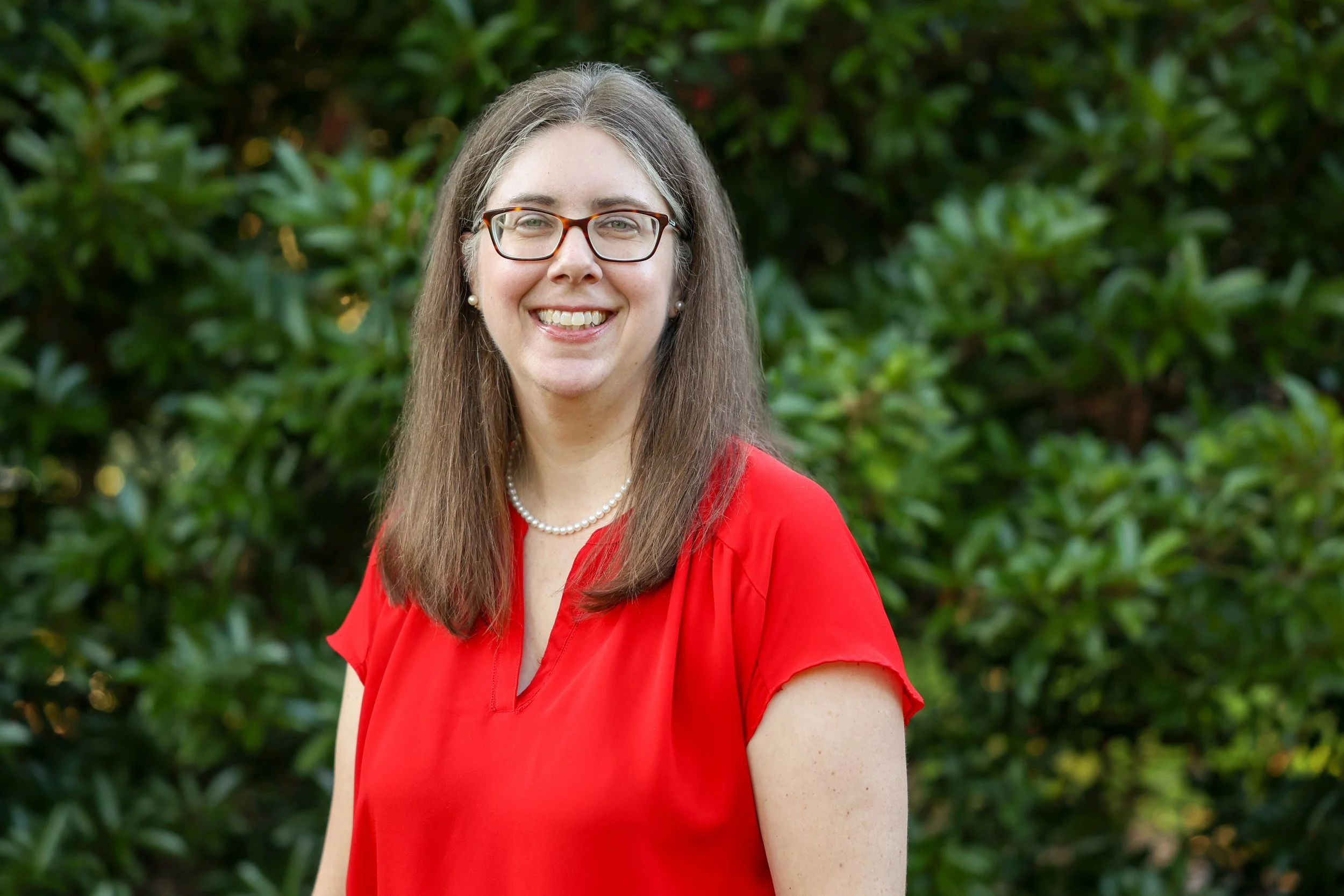 A woman with long brown hair, glasses, and earrings, smiling, wearing a red blouse, standing outdoors with green foliage in the background.