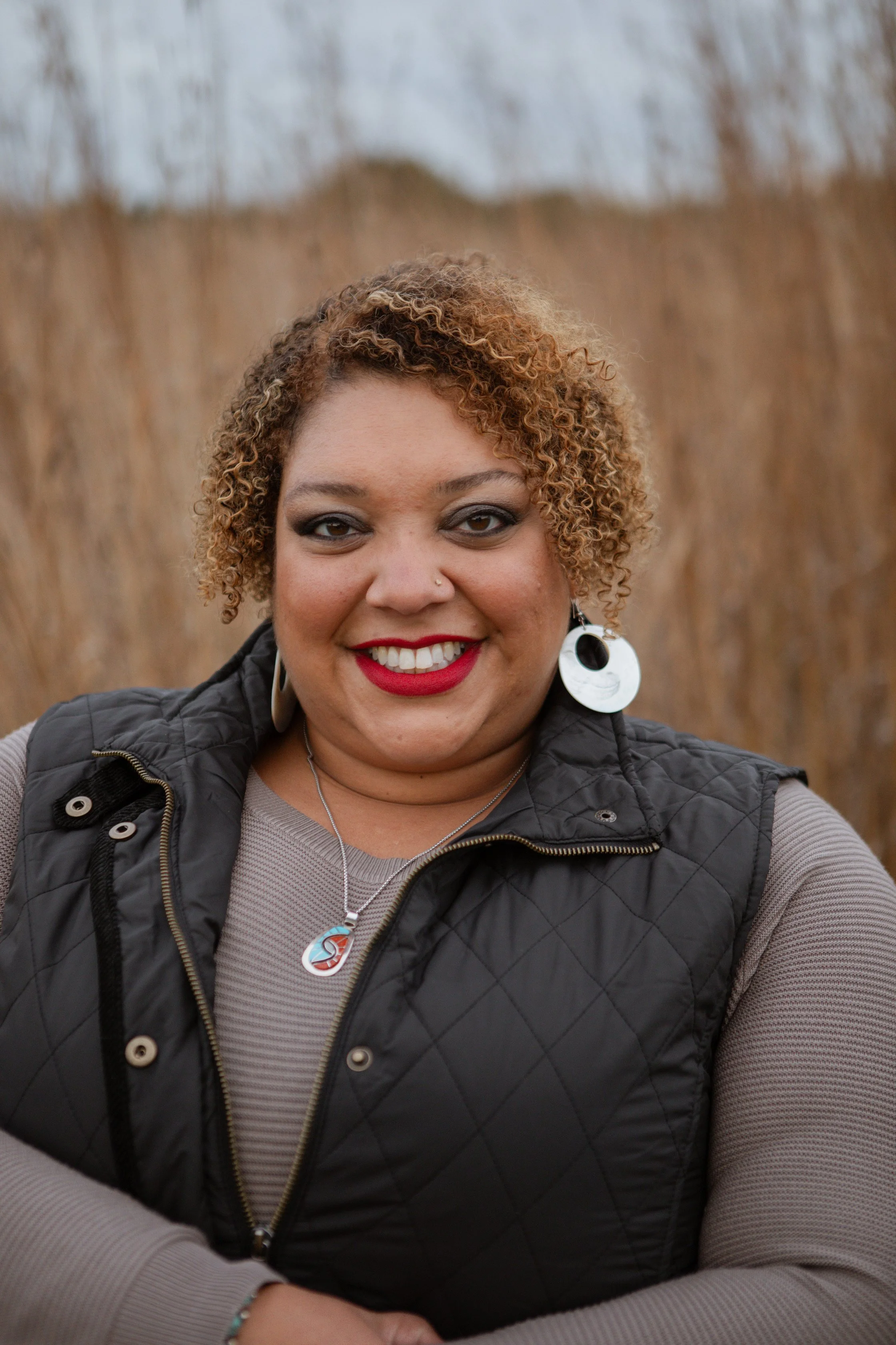 Maddy Day is standing in a brown field. She is smiling at the camera and wearing silver earrings and necklace, with a grey long sleeved shirt with a sleeveless puffy jacket
