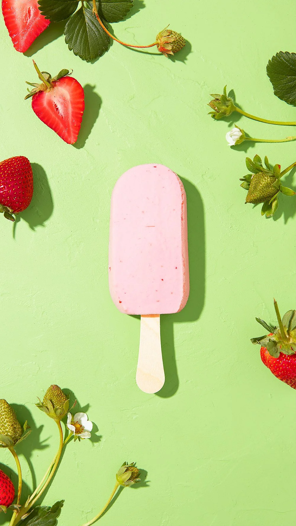 A pink strawberry popsicle on a wooden stick centered on a light green background, surrounded by fresh red strawberries, green leaves, and flower buds.