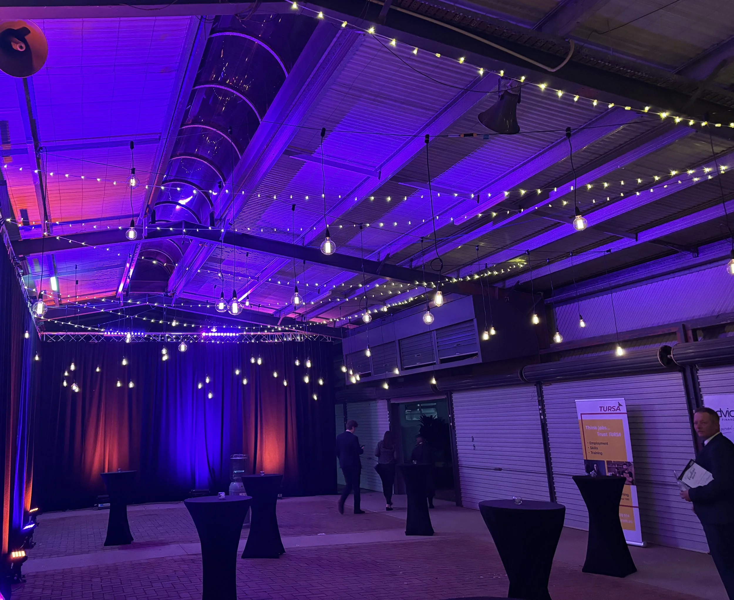 Indoor event space with a stage, purple and orange lighting, hanging string lights, and tall cocktail tables.