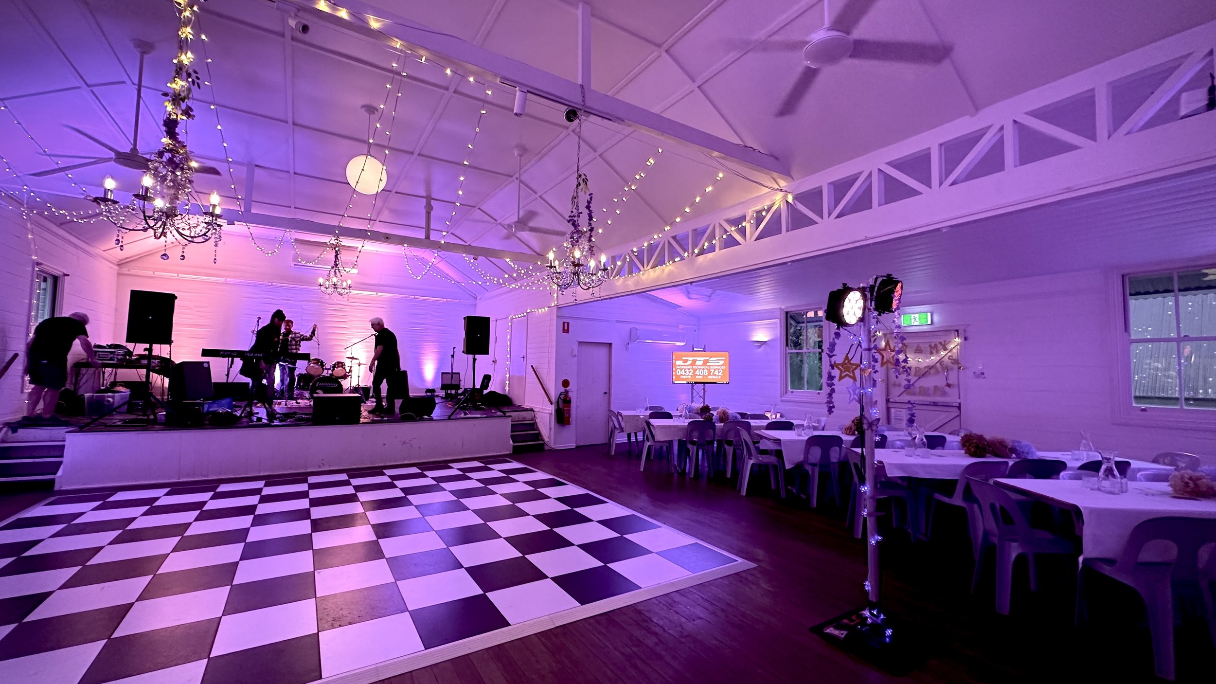 Indoor event space decorated with string lights and chandeliers, stage with musicians preparing to perform, checkered dance floor, and tables set for dining with centerpieces, purple and pink lighting.