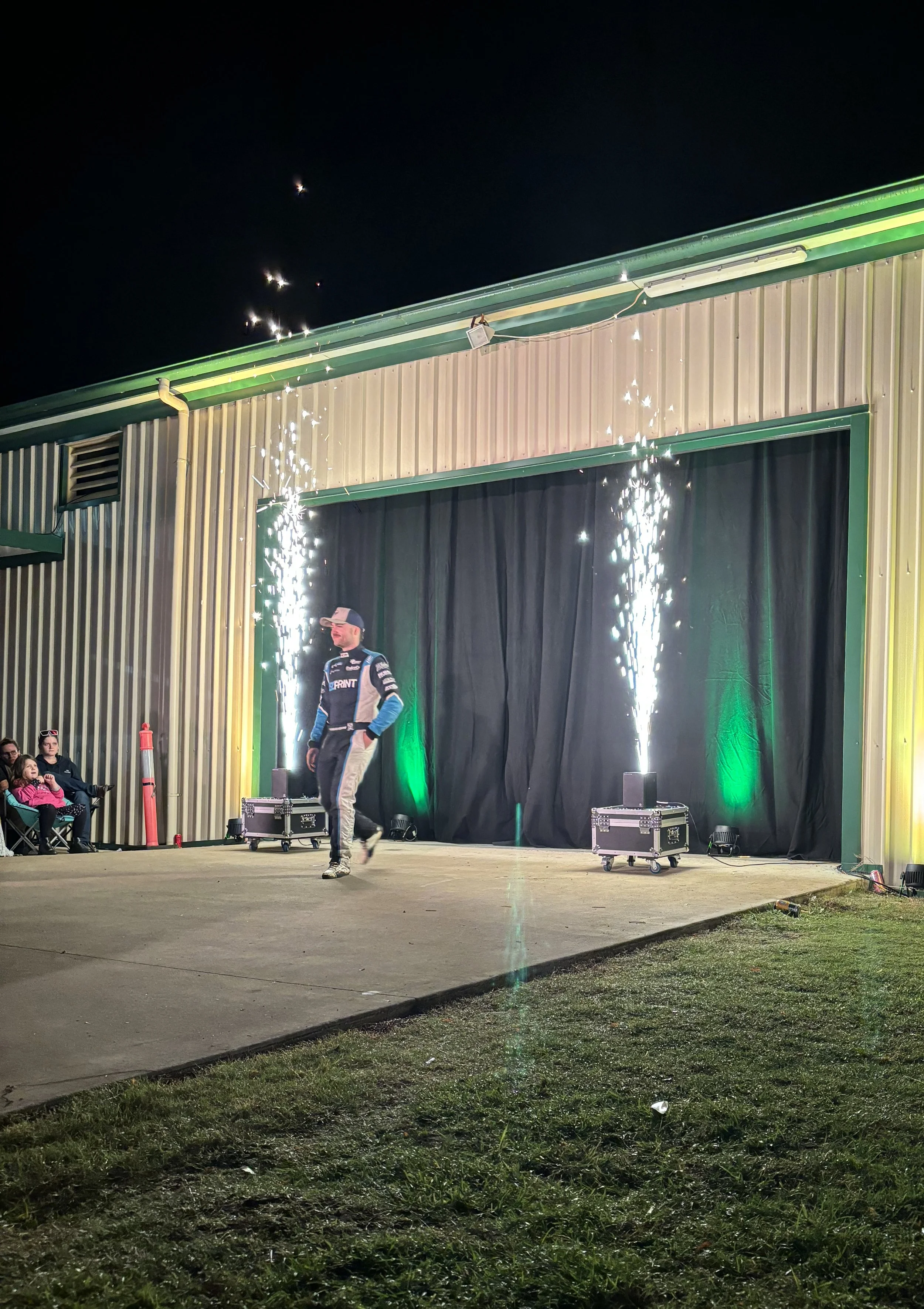 A person walking on an outdoor stage at night with sparklers or pyrotechnics behind them, some audience members seated to the left, and a black curtain backdrop.
