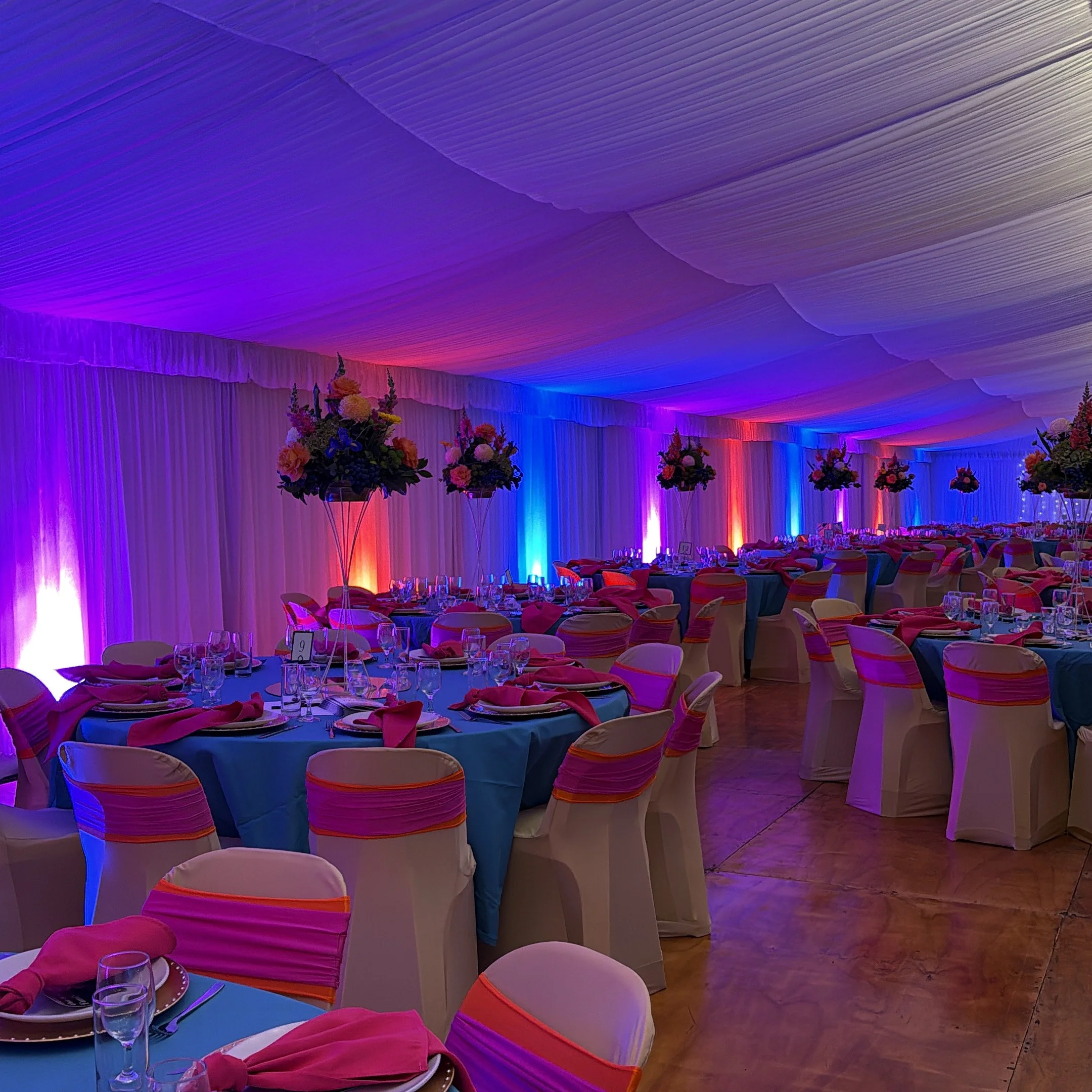 Elegant event space decorated with colorful lighting, floral centerpieces, and round tables set with pink napkins and glassware for a formal gathering.