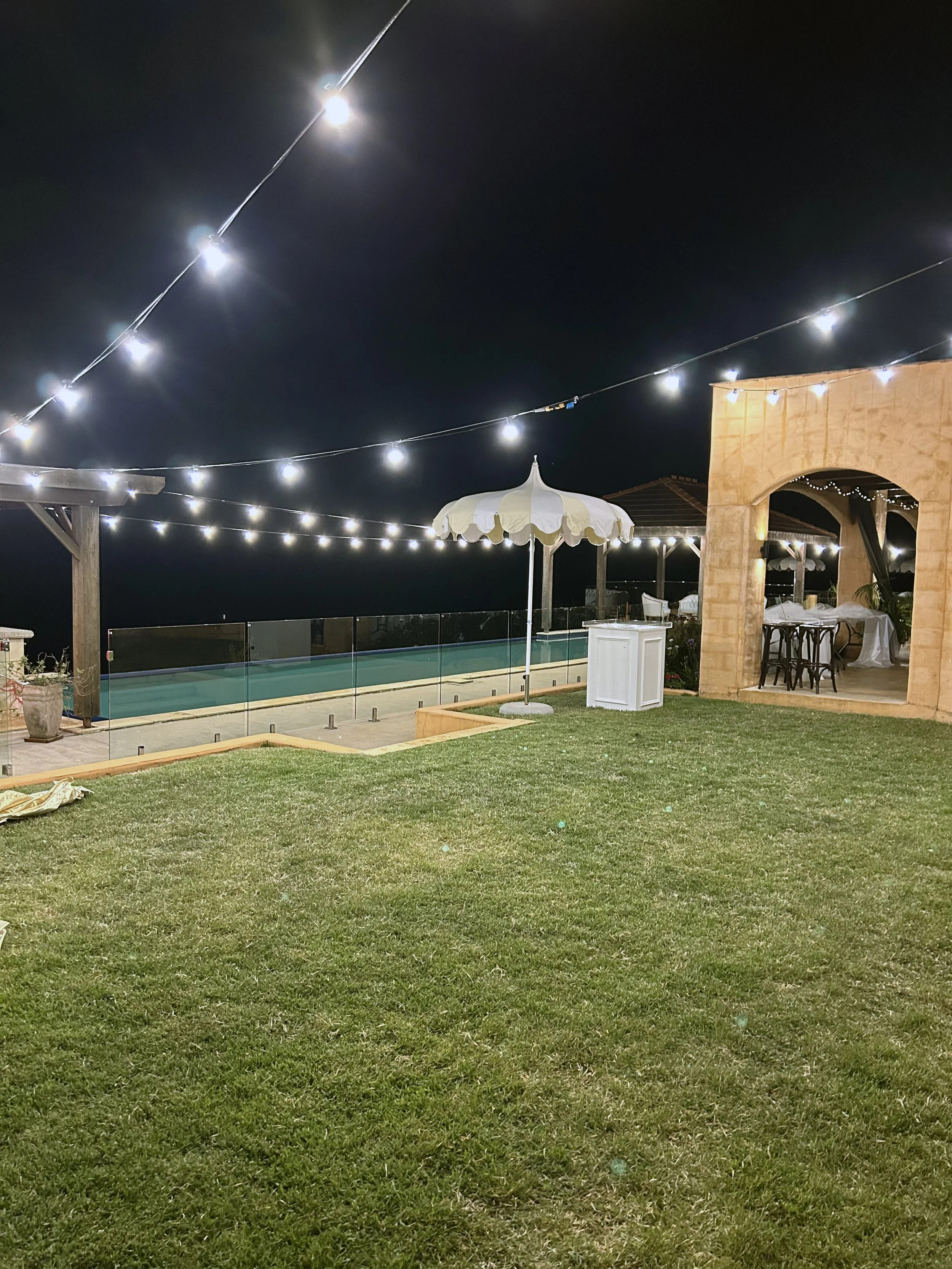 Nighttime outdoor patio with string lights overhead, a swimming pool with a glass barrier, and a beige stucco building with outdoor furniture and an umbrella.