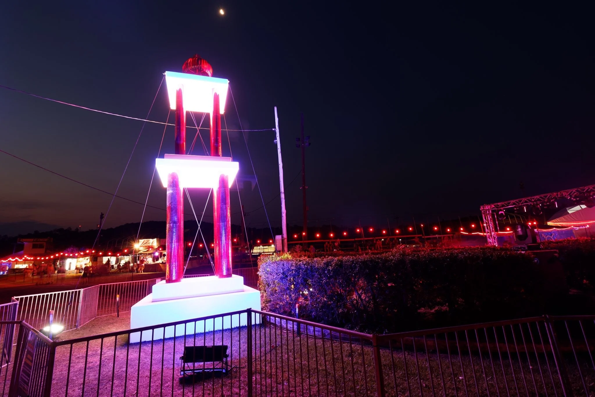 Nighttime view of a neon-lit carnival or fair with a tall illuminated structure, possibly a water tower, decorated with red neon lights, surrounded by fences and carnival rides, with string lights and tents in the background.