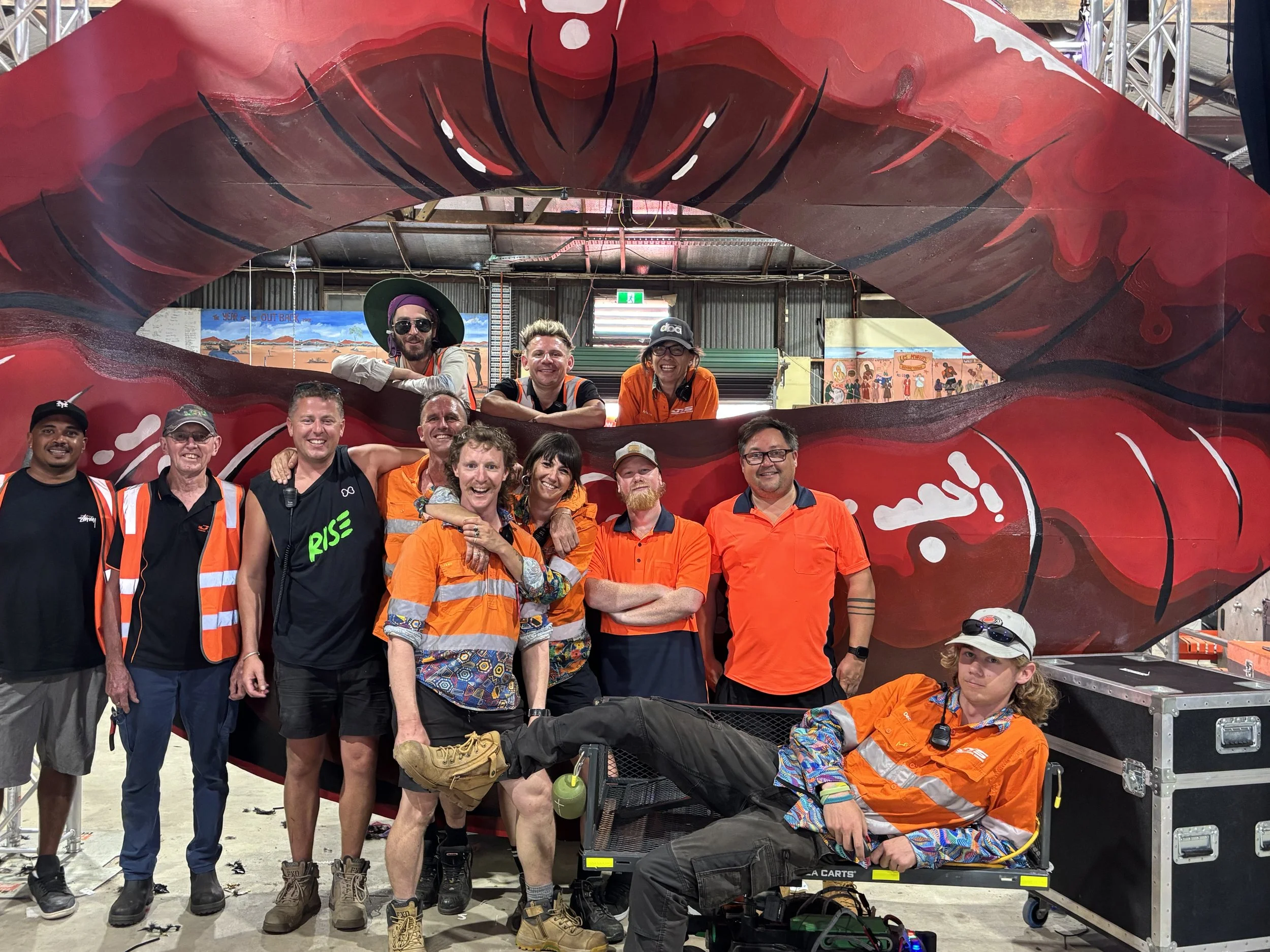 A group of people poses in front of a large structure resembling red lips with an open mouth. Some people are standing, four are wearing orange safety vests, and one person is reclining in a cart. The setting appears to be indoors, possibly at an eve