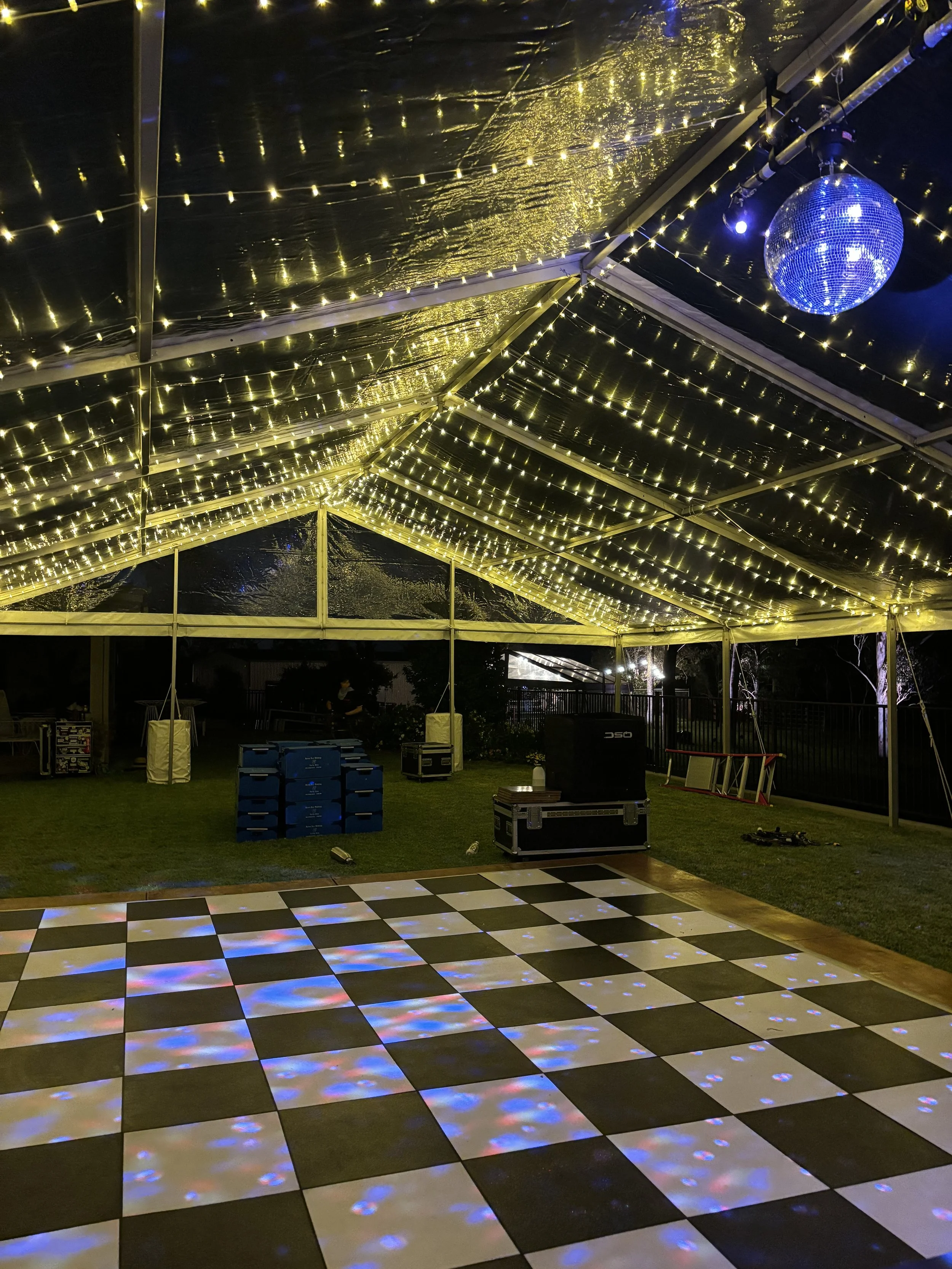 Outdoor dance floor under a transparent tent with string lights and a disco ball; checkerboard pattern on the floor reflecting colorful lights.