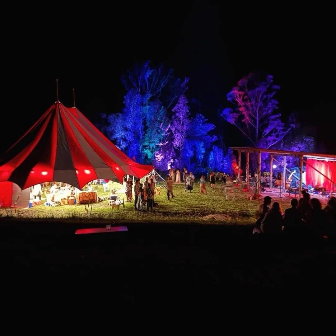 Nighttime outdoor festival scene with illuminated red and white tent, colorful lit trees, and people socializing.