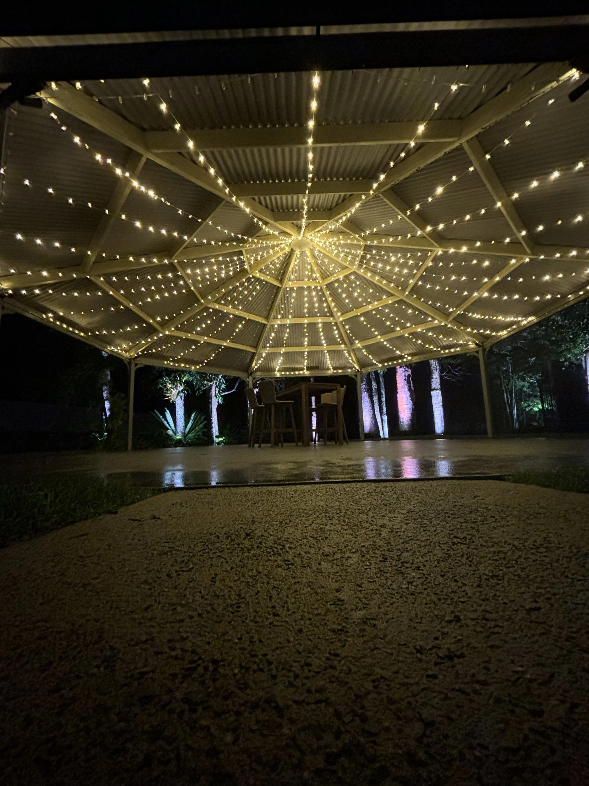 Outdoor gazebo at night with string lights under the roof, illuminating the space. A few chairs are visible in the background. The ground is lit with a spotlight on some trees in the distance.