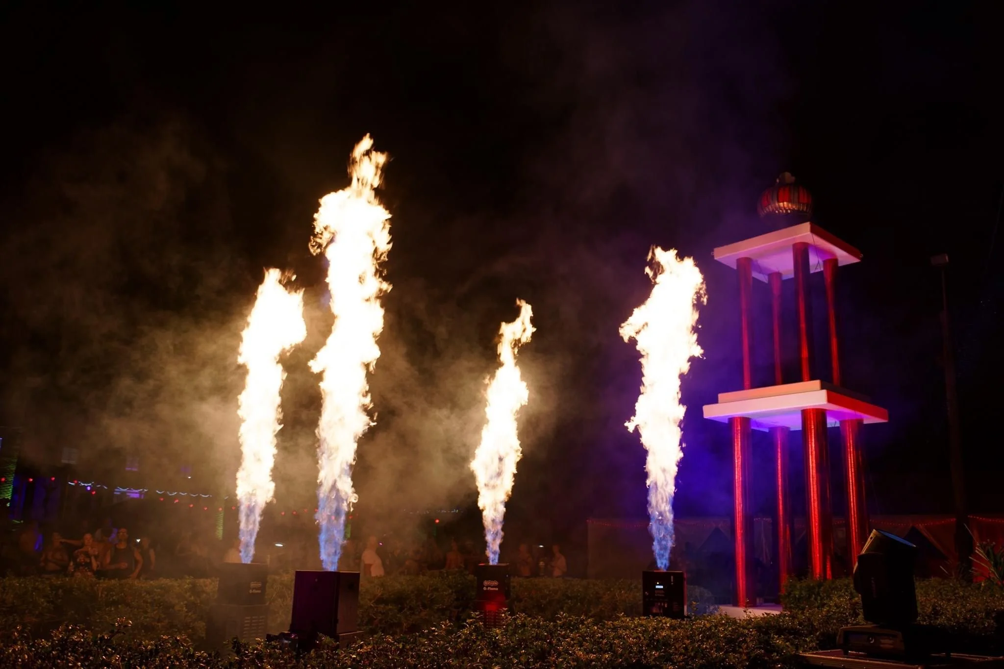 Nighttime outdoor fire performance with flames shooting upward from multiple devices, and a lit-up stage-like structure with purple lighting.