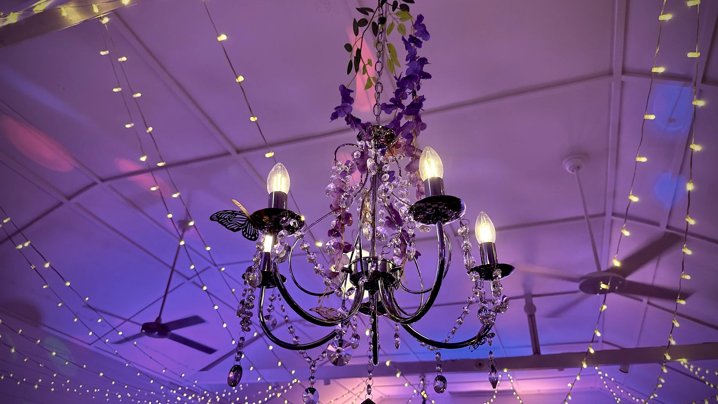 Chandelier decorated with crystals and butterflies hanging from the ceiling, surrounded by purple, yellow, and pink lighting, with ceiling fans in the background.