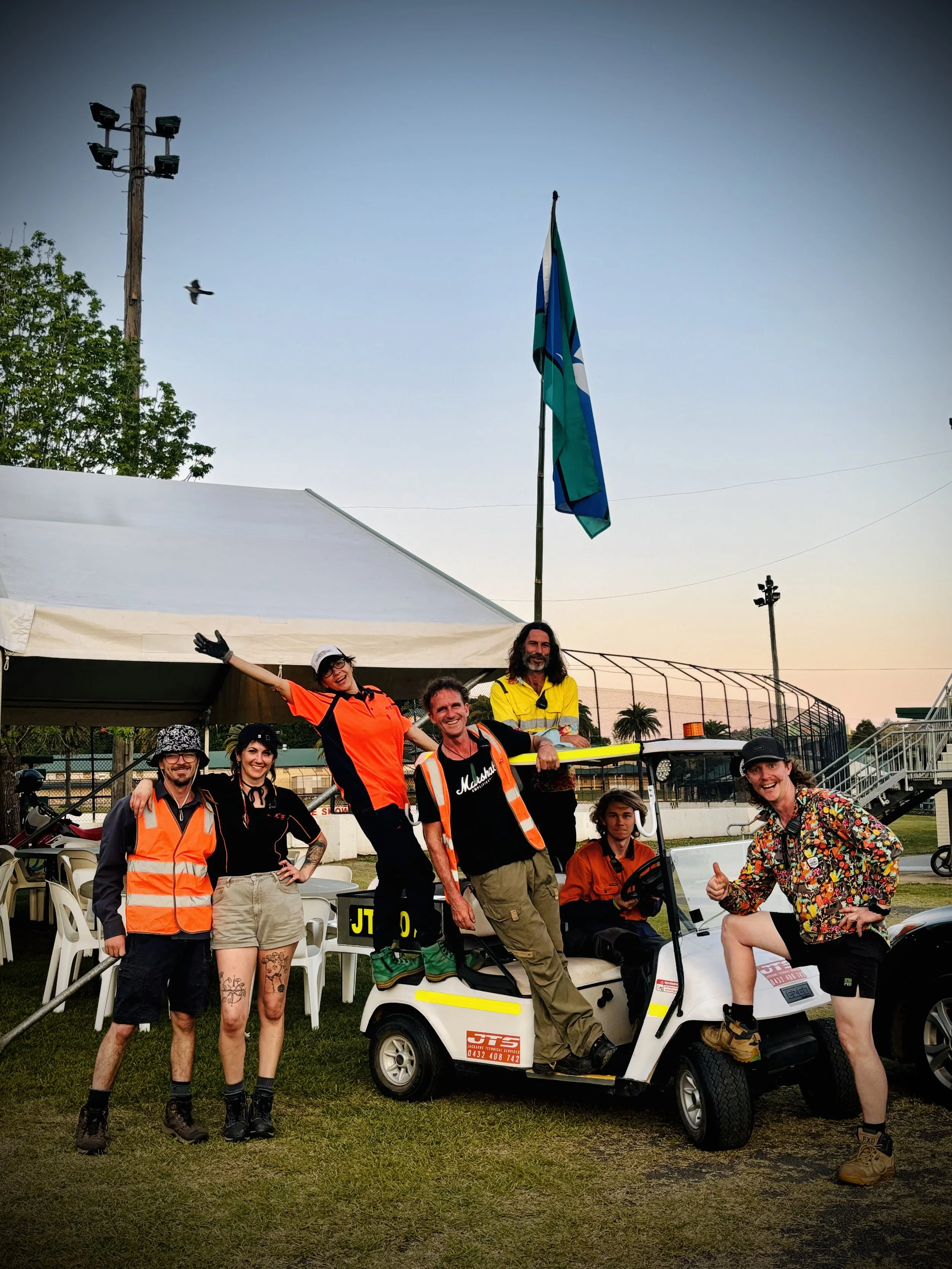 Group of people in casual and safety gear posing with a golf cart at an outdoor event, with tents and a flag visible in the background.