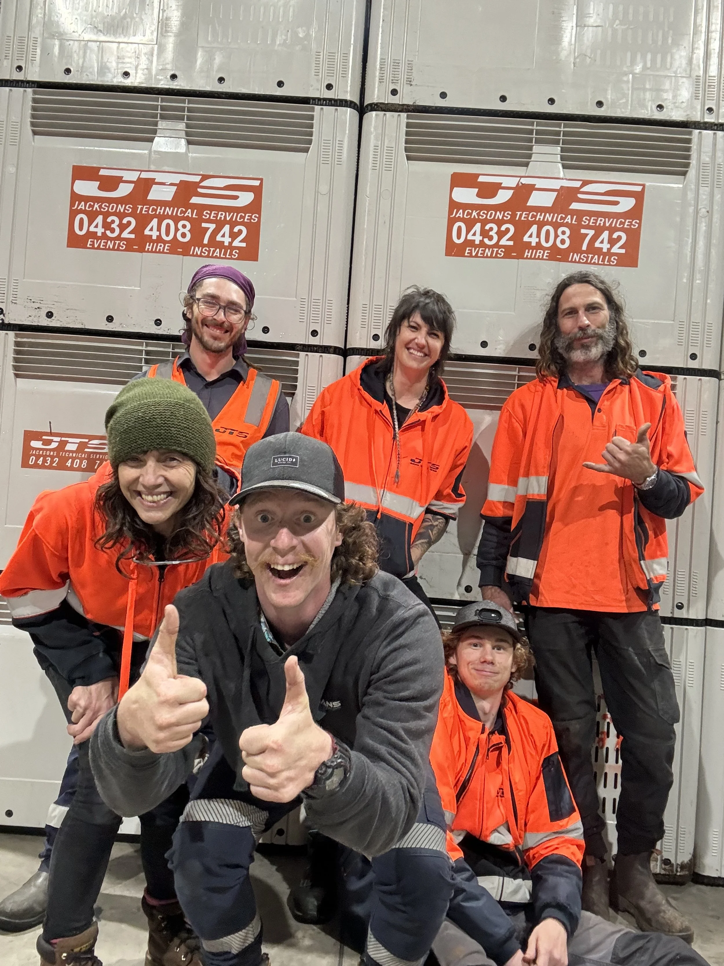 Group of six people wearing orange work vests in front of "Jacksons Technical Services" sign.