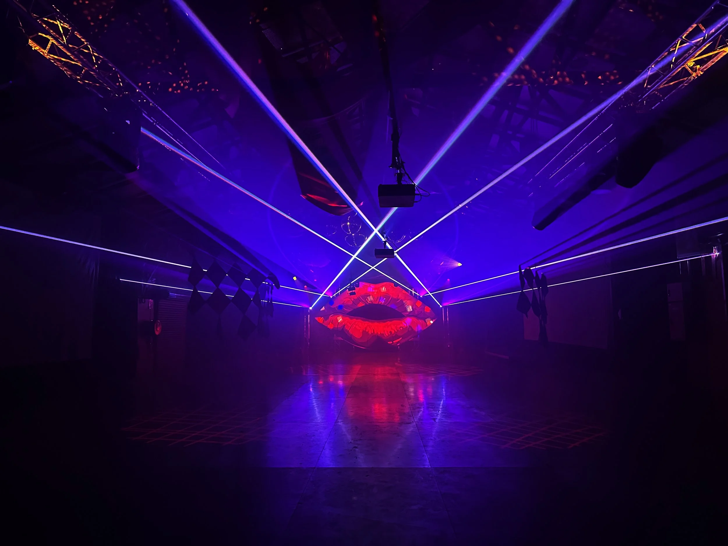 Dark nightclub with purple and blue laser lights, a large neon red lips sign at the back, and a mirrored floor reflecting the lights.