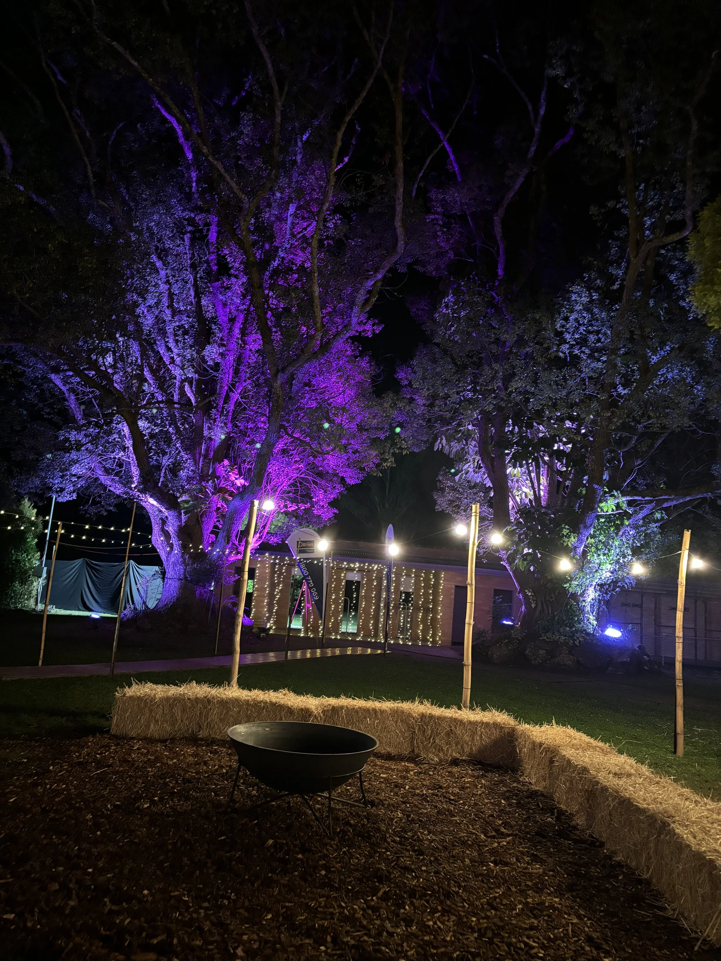 A nighttime outdoor scene featuring trees illuminated by colorful lights, with string lights and a structure in the background. Hay bales form a seating area around a fire pit on a grassy area.
