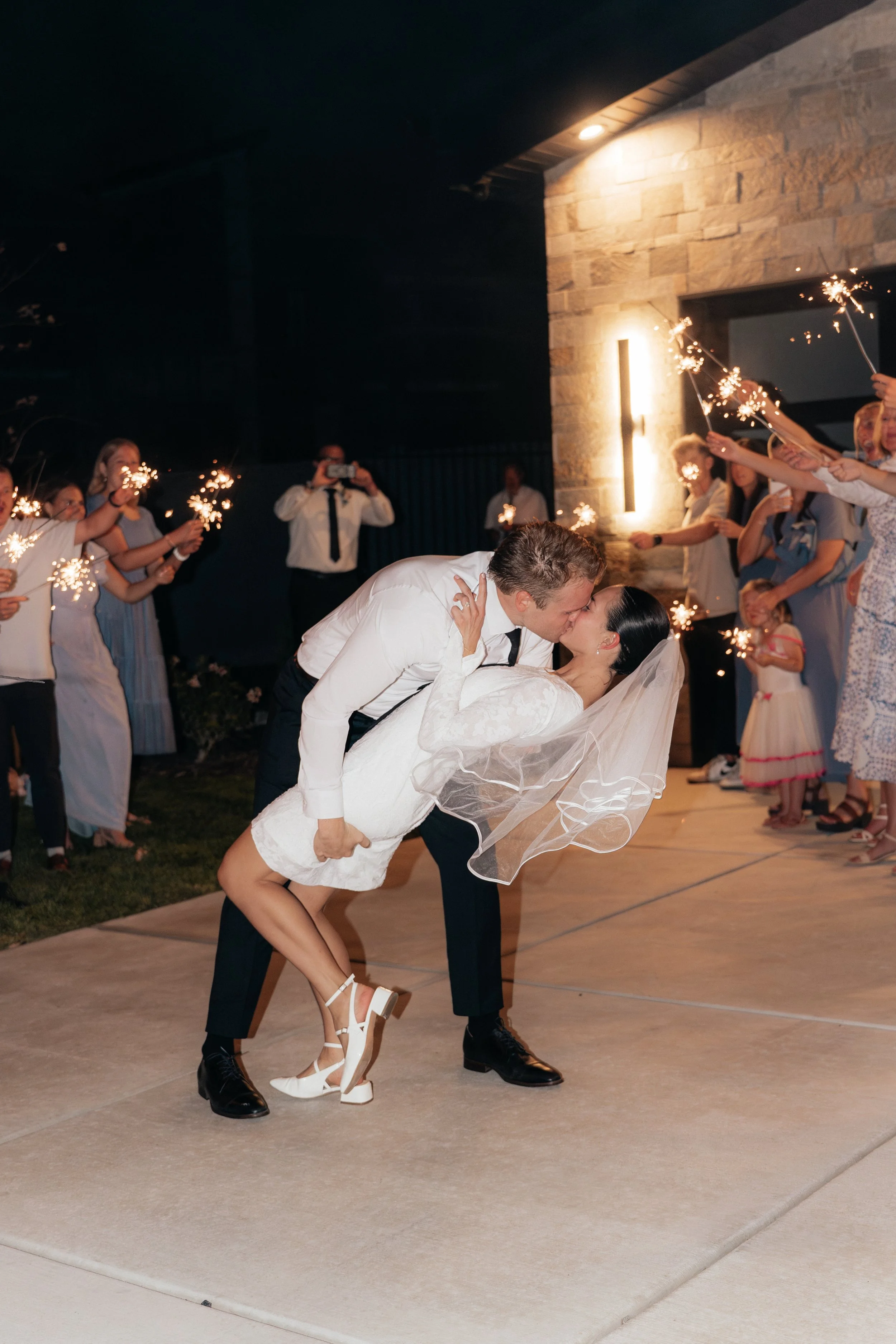 A bride and groom holding hands and smiling after their wedding ceremony outdoors by a lake, with mountains and cloudy sky in the background, surrounded by seated guests and wedding party.