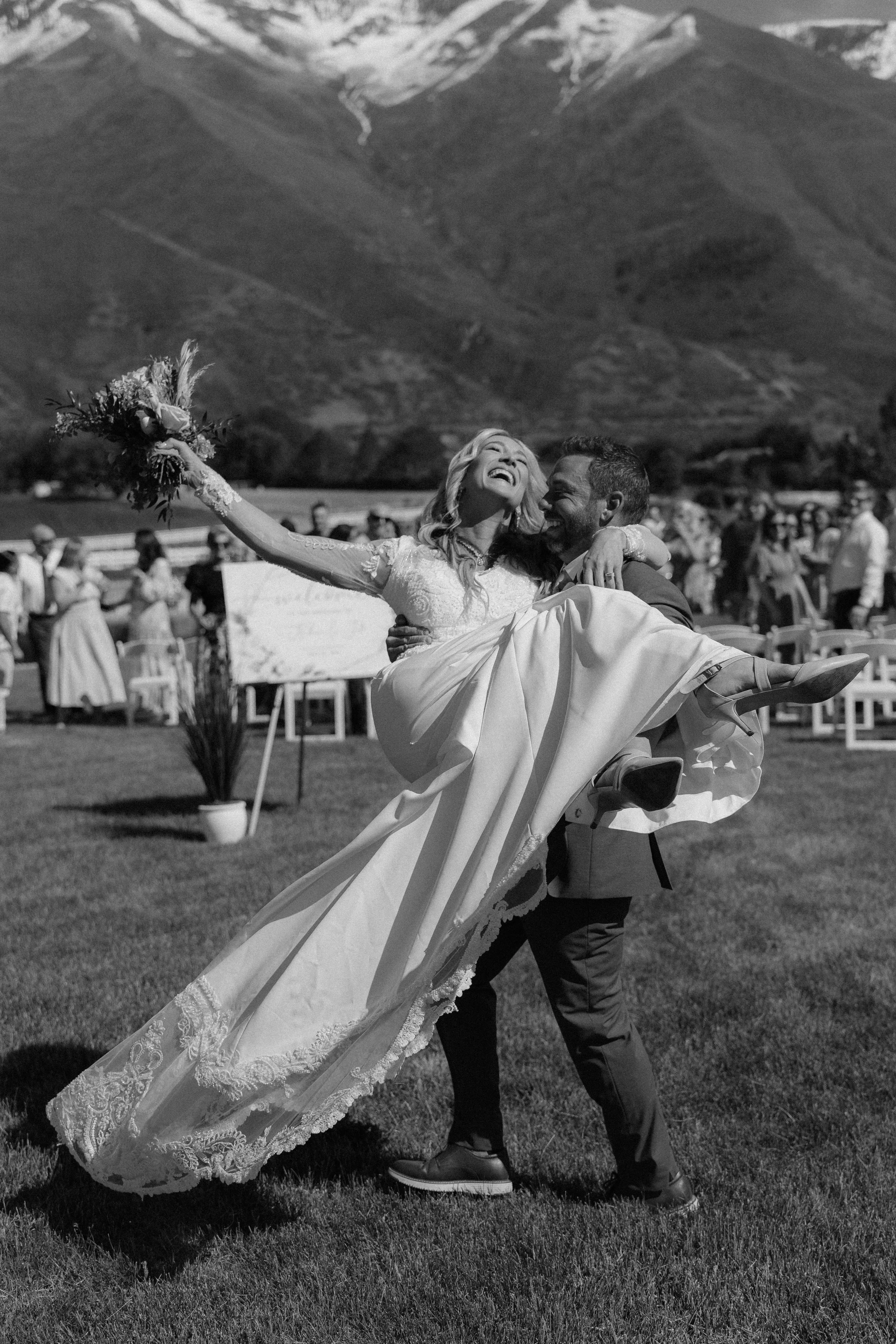 A bride and groom holding hands and smiling after their wedding ceremony outdoors by a lake, with mountains and cloudy sky in the background, surrounded by seated guests and wedding party.