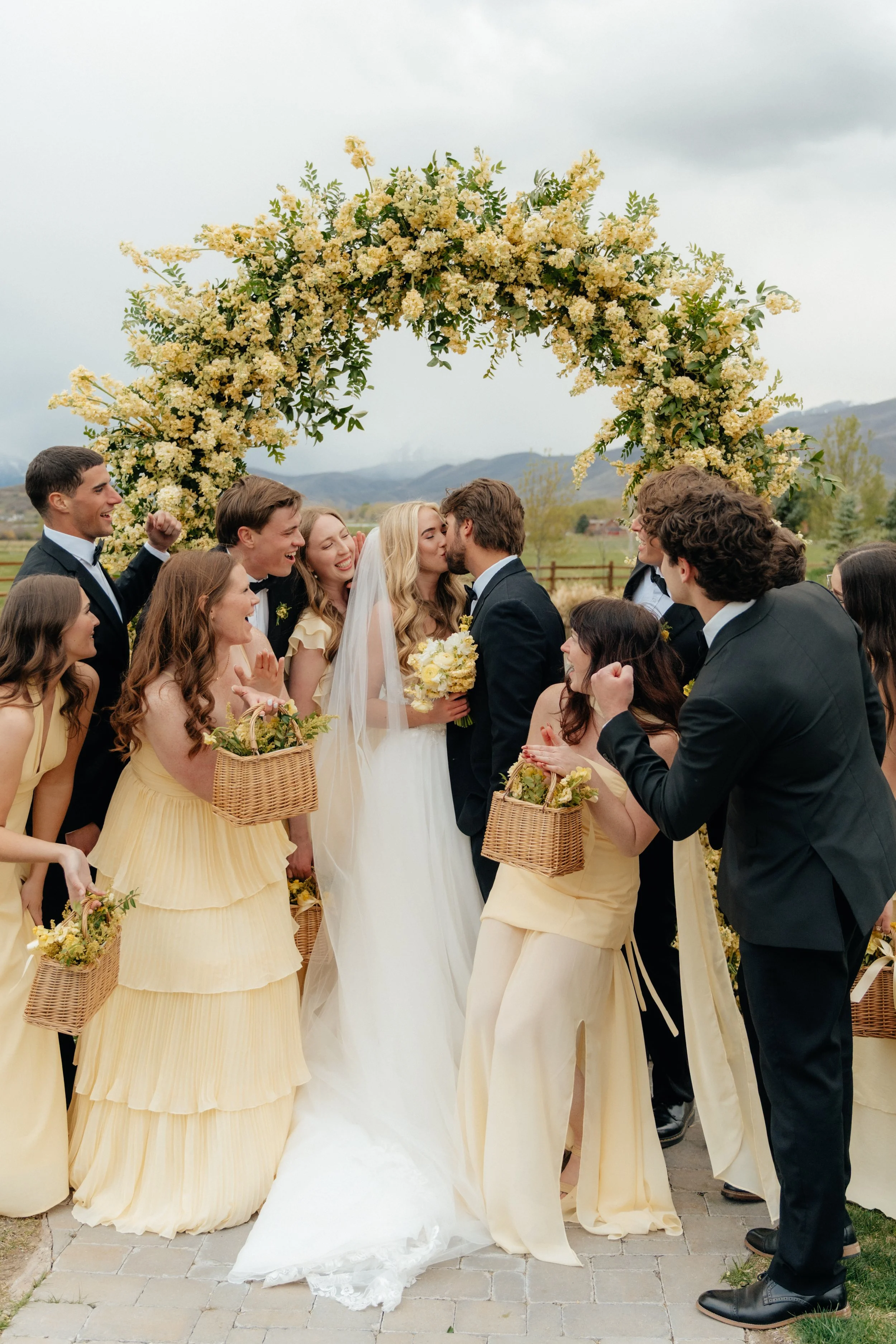 A bride and groom holding hands and smiling after their wedding ceremony outdoors by a lake, with mountains and cloudy sky in the background, surrounded by seated guests and wedding party.