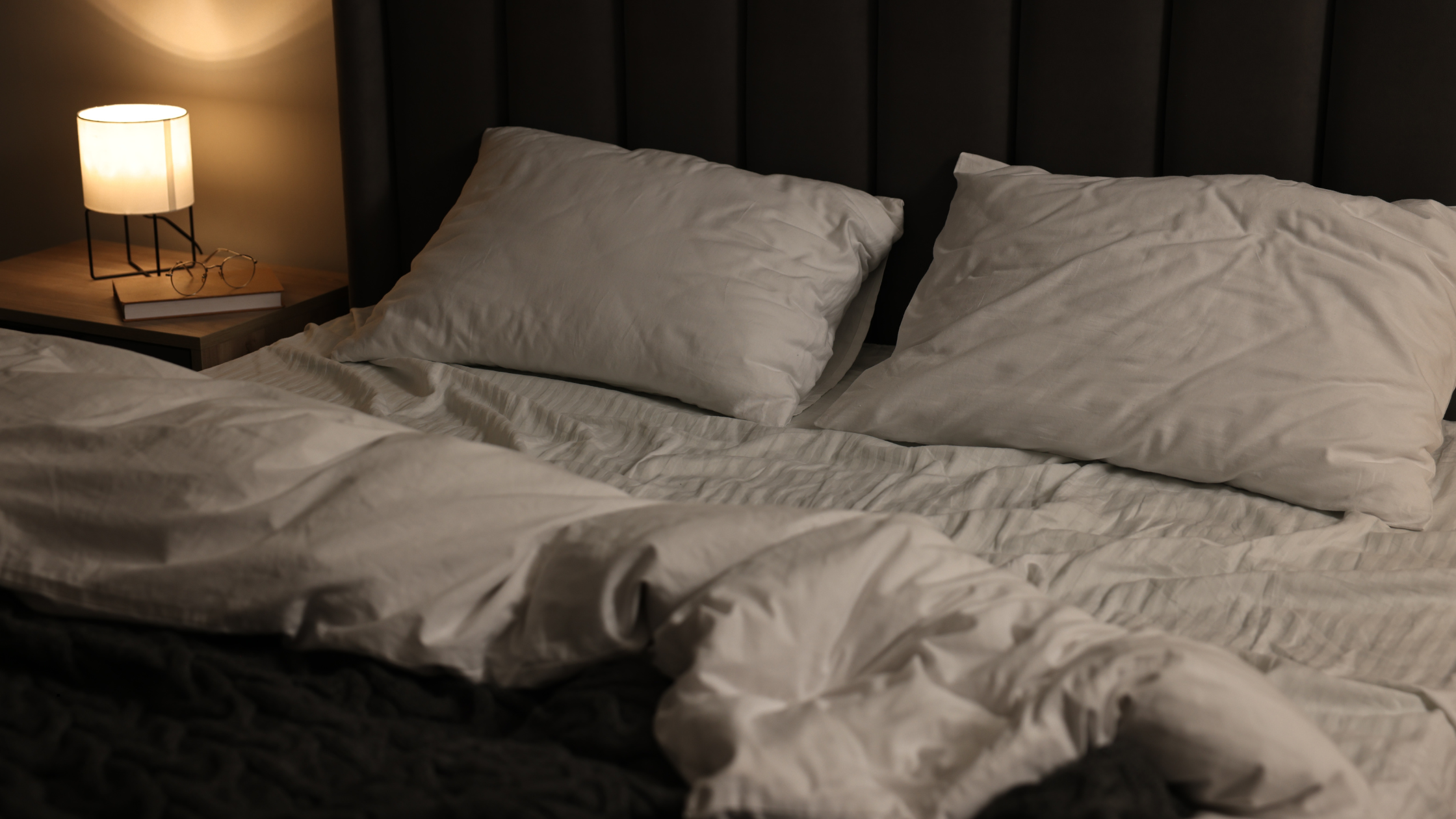 A softly lit bedroom at night featuring an unmade bed with white linen, dark upholstered headboard, and a warm bedside lamp casting a gentle glow over a stack of books and glasses on the nightstand.