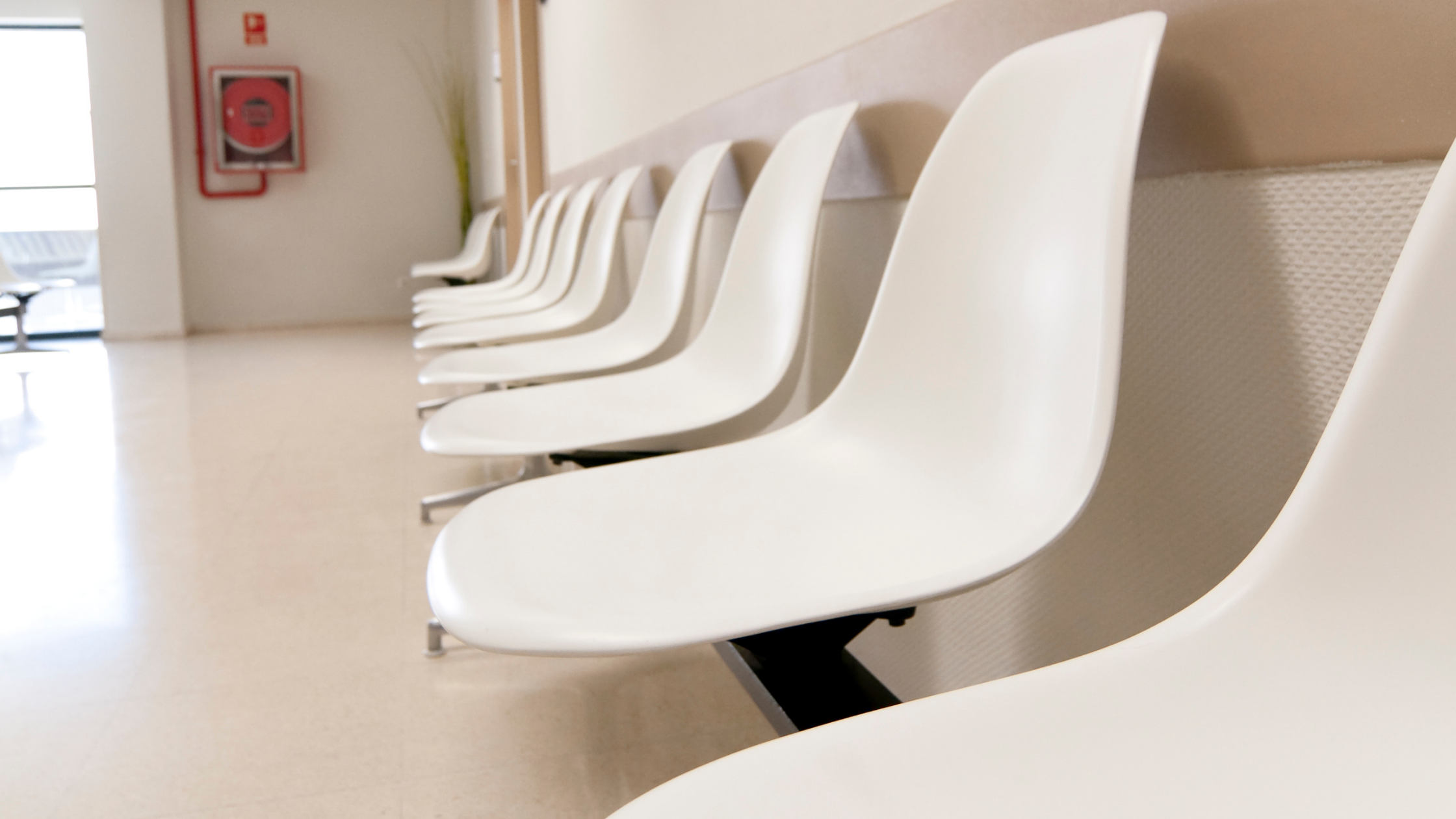 Empty row of identical white plastic chairs in a clinical waiting room, photographed from a low angle along a pale featureless wall, with a fire extinguisher sign and a partially visible corridor in the background.