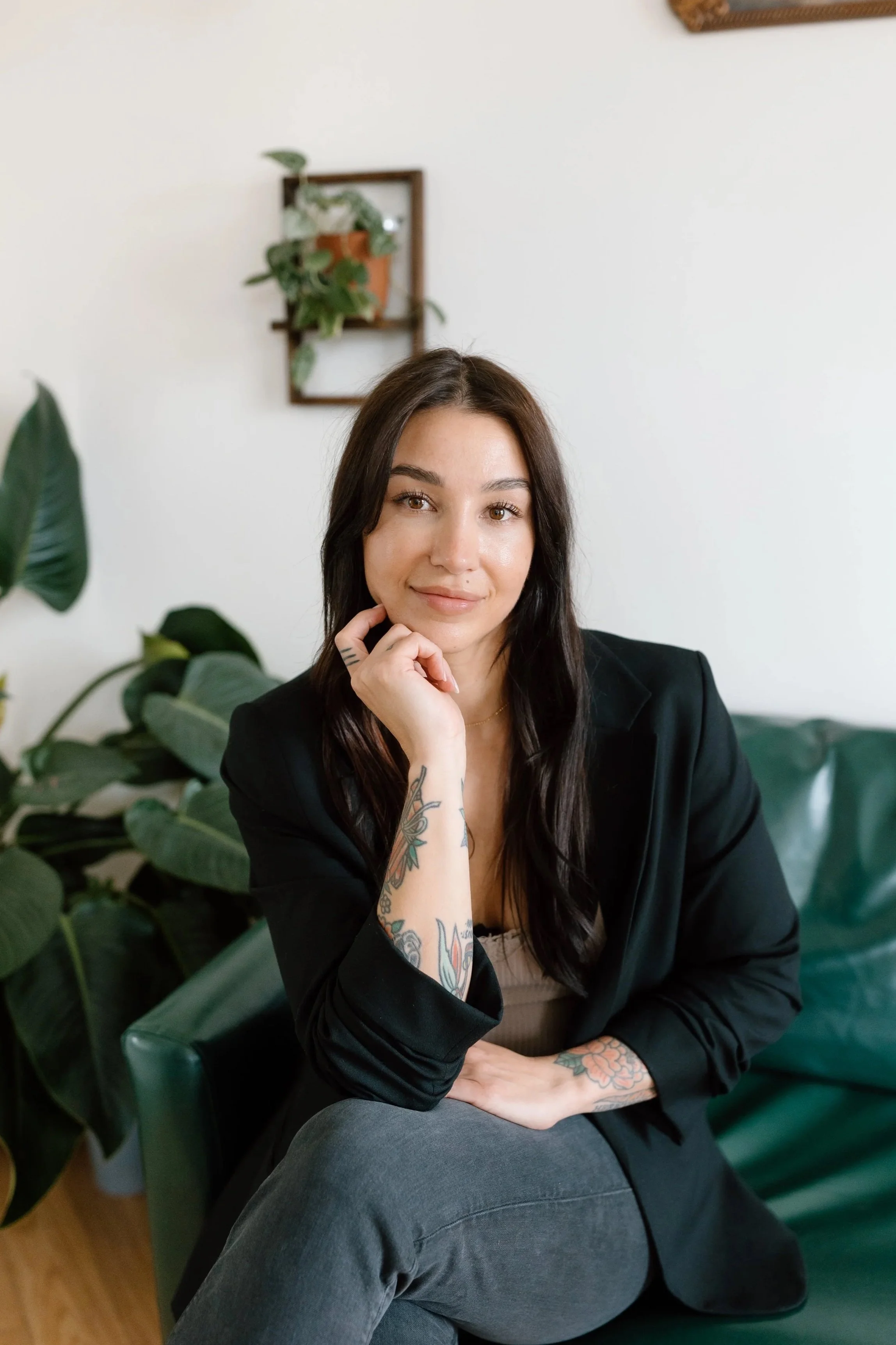 A woman with long dark hair, wearing a beige sweater and blue jeans, standing indoors with her arms crossed, smiling at the camera. The background features white walls decorated with hanging plants, wooden shelves with glass jars, and a framed picture.