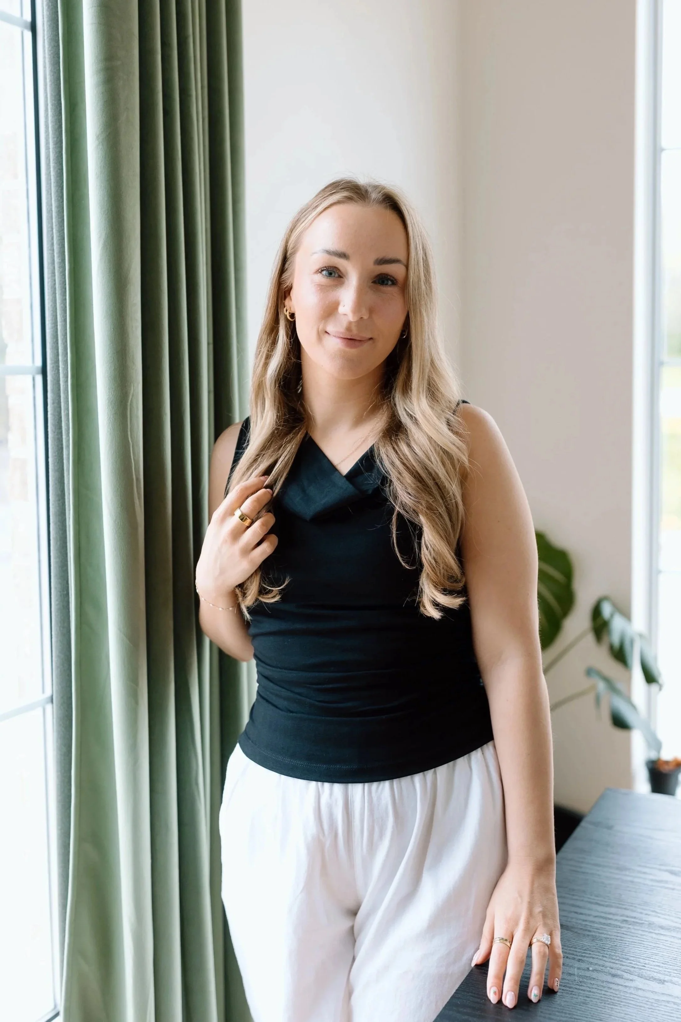 A smiling woman with long blonde hair standing indoors in front of a decorative wooden mirror and green plants, dressed in a black shirt and black pants.