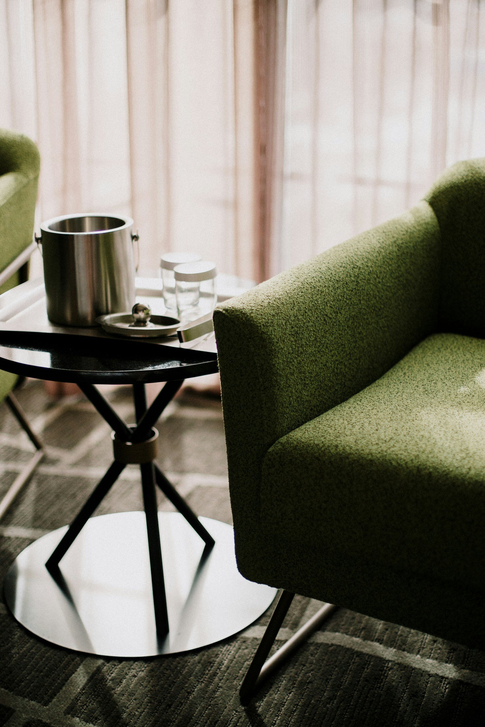 A side view of a small round black table with a white base, holding a stainless steel ice bucket, two glasses, and a small silver object. Next to the table is a green upholstered armchair, all positioned on a patterned rug in front of sheer curtains.