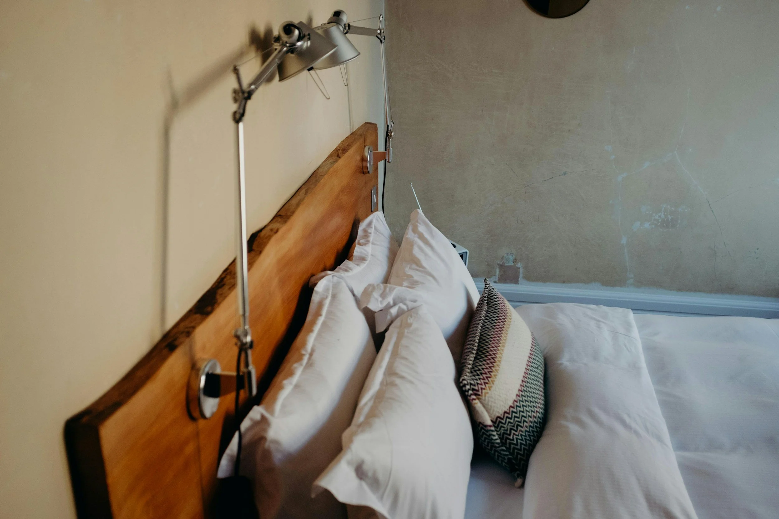 A bedroom with a wooden headboard, white bed linens, pillows, and a patterned throw blanket. A wall-mounted light fixture is above the headboard, and the wall behind has a distressed, textured appearance.