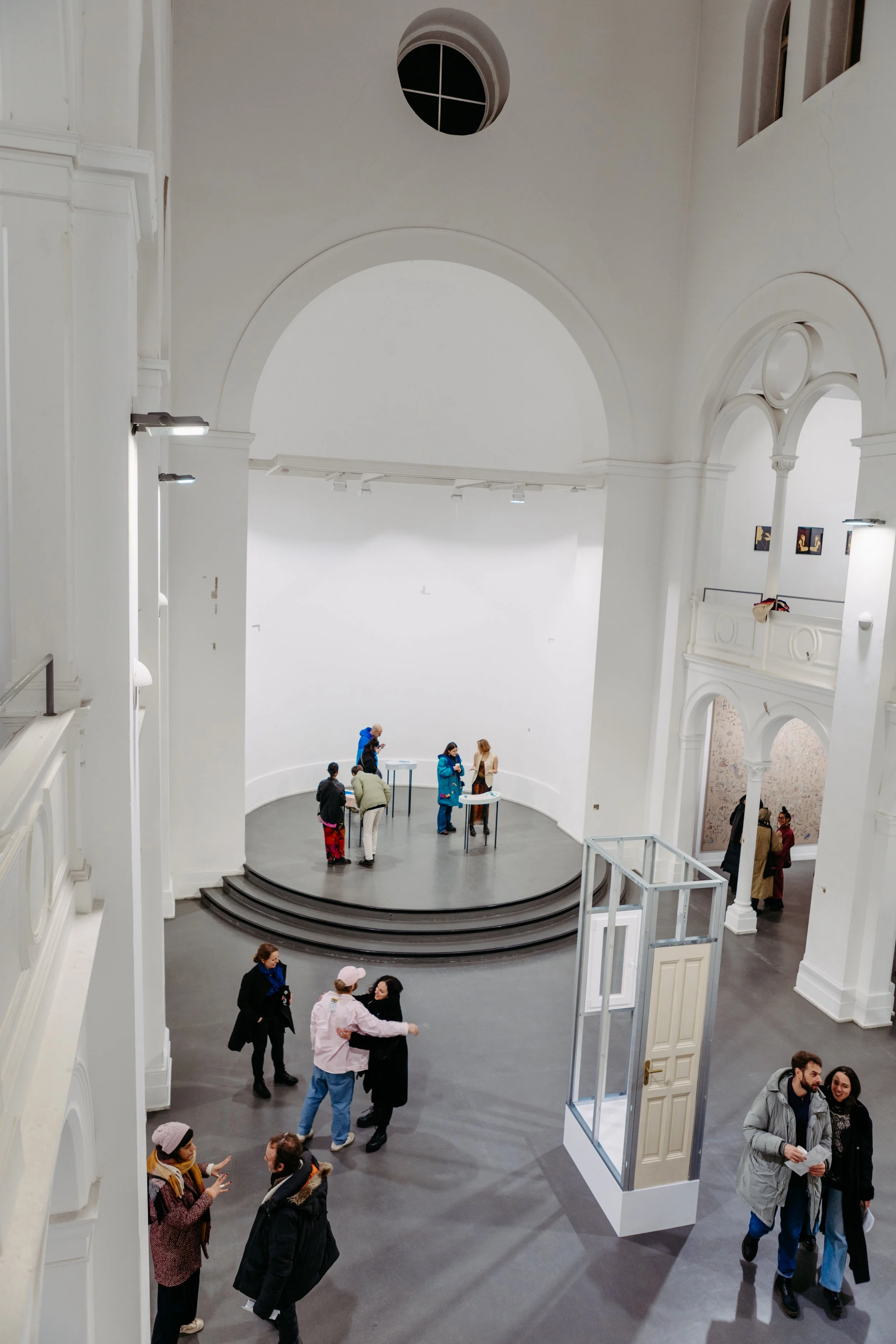 Interior of a museum or gallery with visitors walking and conversing. The space has white walls, arched windows, and a staircase. People are gathered in groups, some near small tables, and some near the doors.