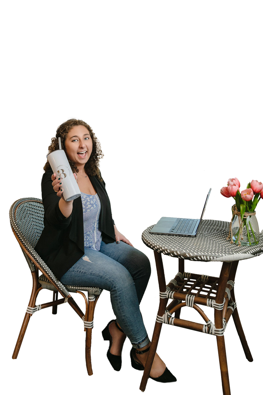 Owner and graphic designer behind Brandelle Designs sitting at a wicker table with a laptop, holding a white water bottle, smiling, with a bouquet of pink tulips on the table.