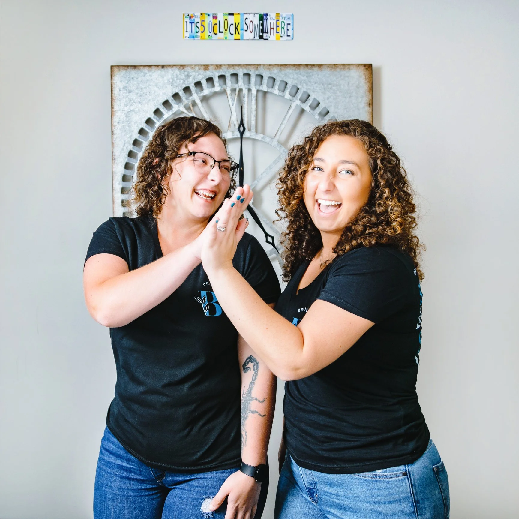 Two women smiling and high-fiving in front of a large clock and a sign that reads 'It's 5 o'clock somewhere'. They are wearing black T-shirts and jeans.