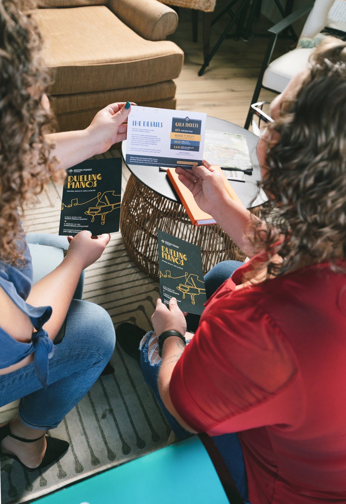Two women sitting on a striped rug with a round coffee table, sharing promotional flyers for a dueling pianos event, while one holds a flyer about gala tickets and the other about the event details.