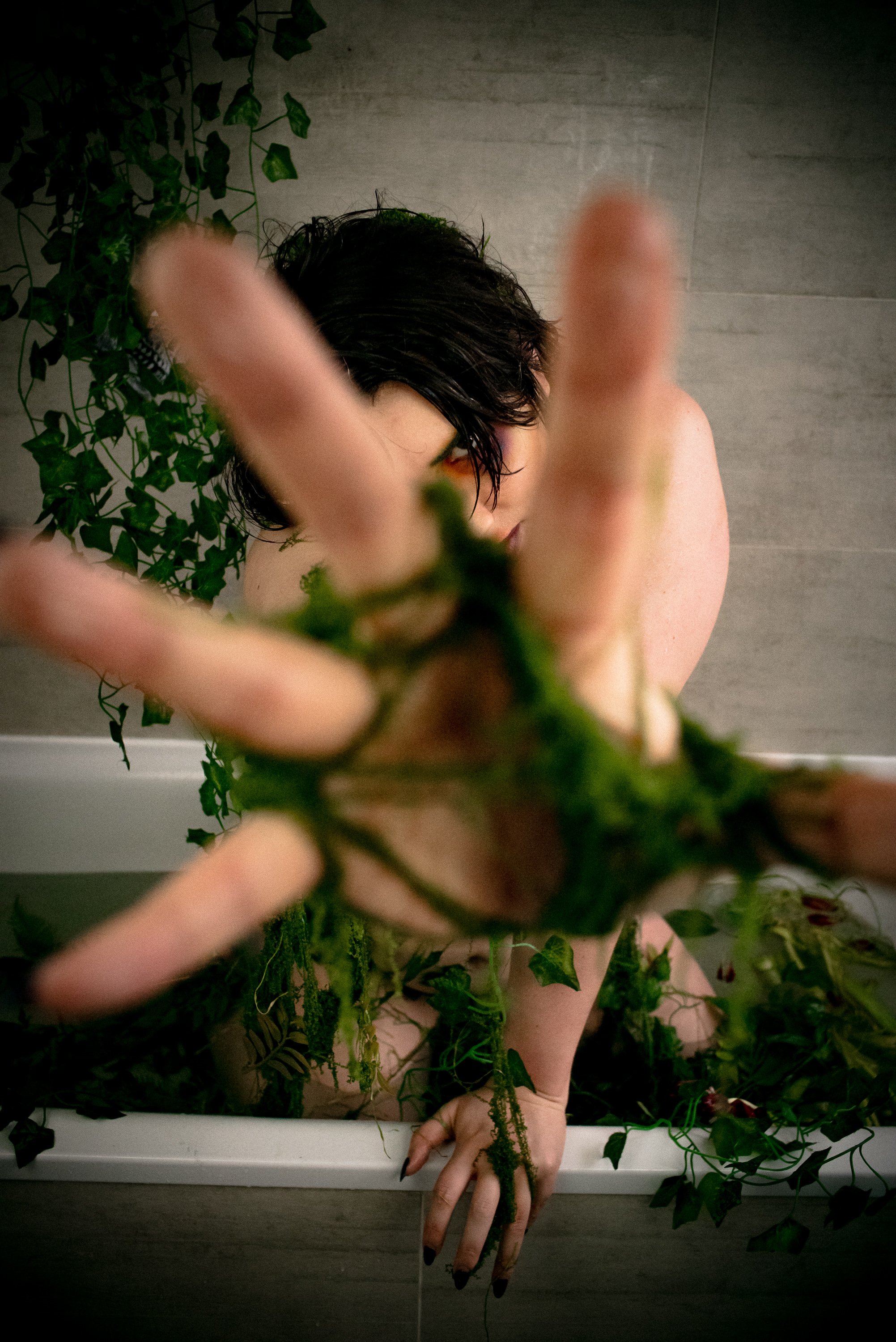 Person with black hair, partially obscured, reaching towards the camera, surrounded by green ivy and plants, in a room with wood-patterned walls.