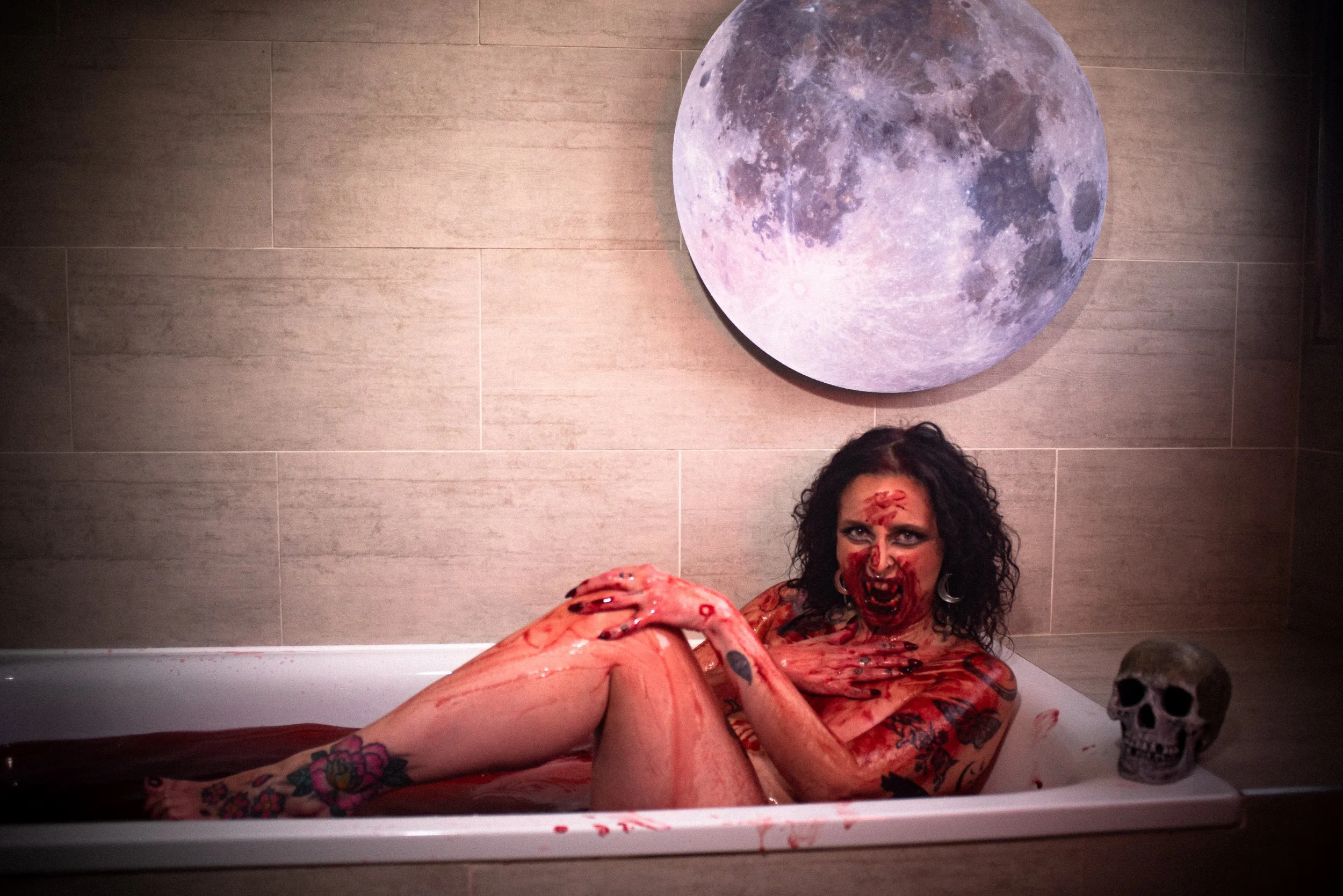 A woman dressed in horror makeup lying in a bloody bathtub next to a skull. There is a large moon decoration on the wall behind her.