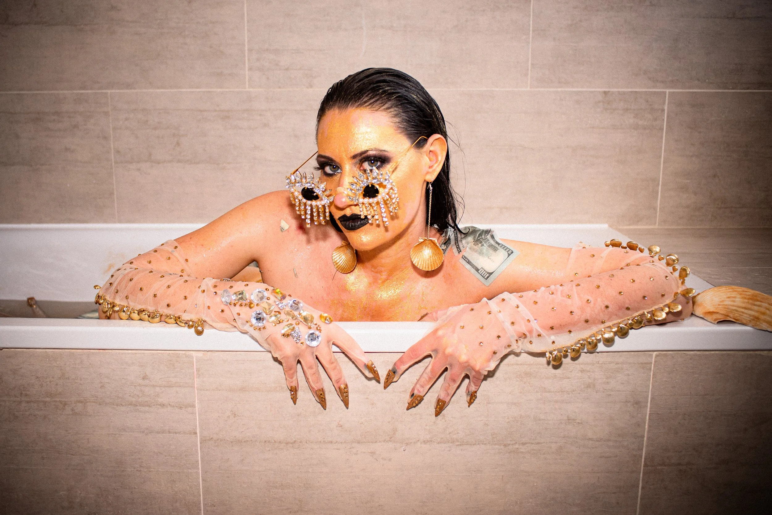Person with dark wet hair and makeup submerged in a bathtub filled with water, adorned with shiny jewelry and dollar bills, with a plain tiled wall in the background.