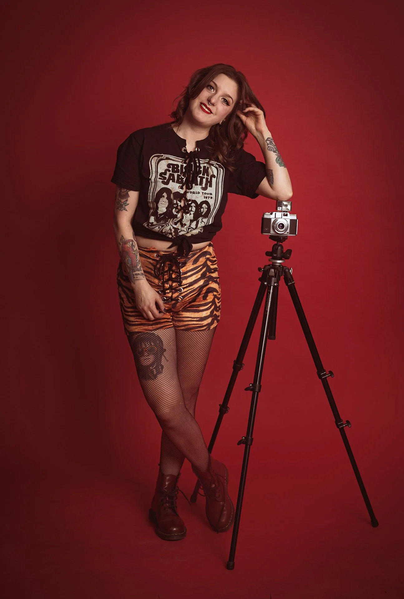 A young woman with brown hair, tattoos, and red lipstick posing next to a vintage camera on a tripod against a red background. She is wearing a black graphic t-shirt, tiger print shorts, fishnet stockings, ankle boots, and is smiling at the camera.