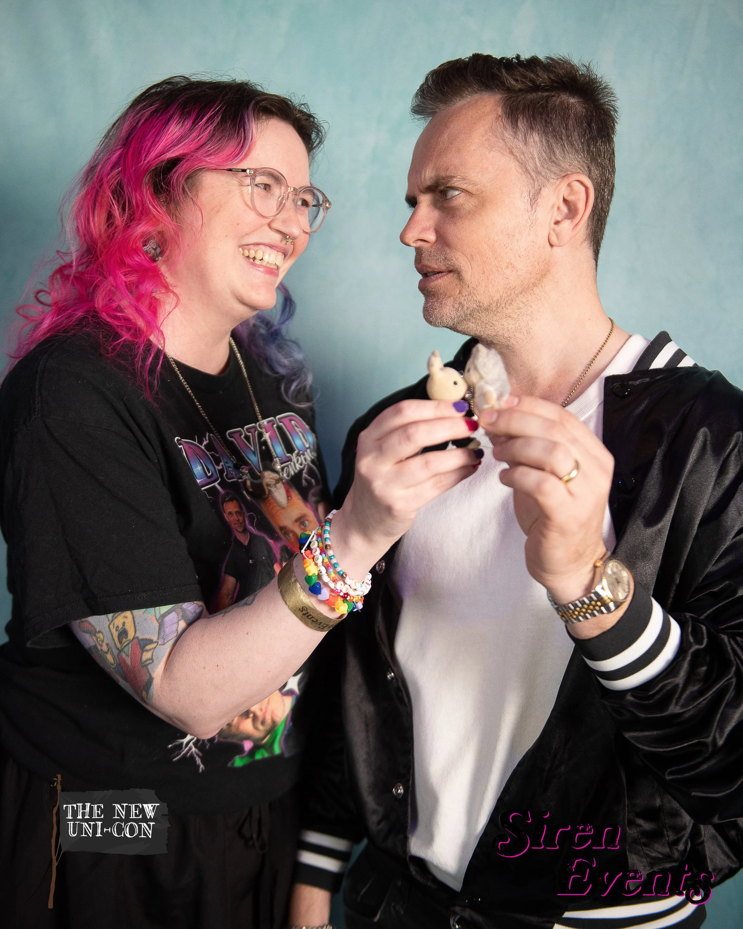 A woman with pink and purple hair smiling and holding a toy pig toward a man with short dark hair who is looking at her. The woman is wearing a black graphic t-shirt and colorful bracelets on her wrist, while the man is wearing a white t-shirt, black jacket, and gold watch. The background is teal. There are logos in the bottom corners: 'THE NEW UNI-CON' on the left and 'Sirén Events' on the right.