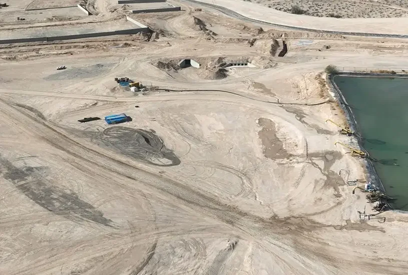 Aerial view of a construction site by a body of water, with heavy machinery and equipment on sandy terrain, and dirt roads crisscrossing the area.