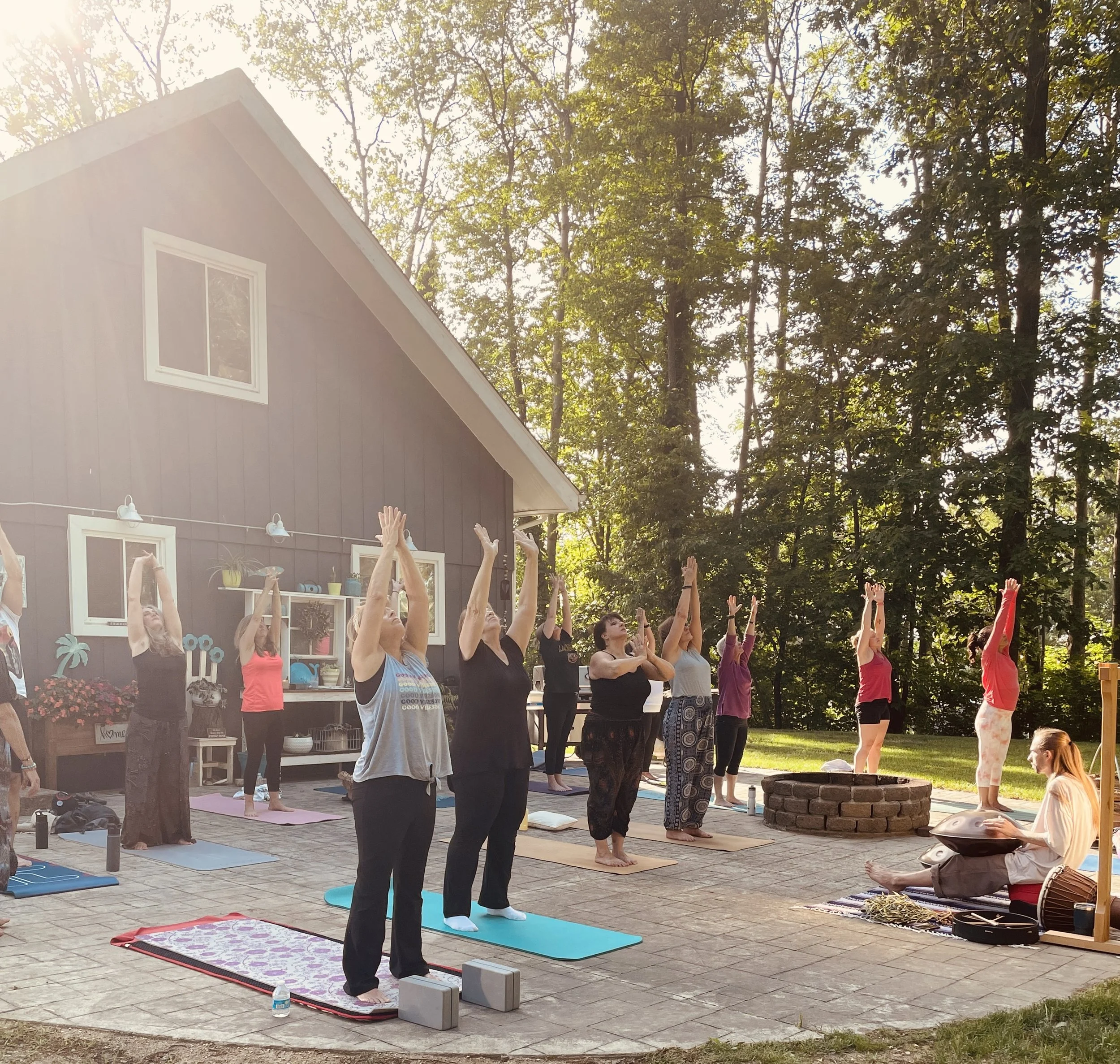 A group of people practicing yoga outdoors on a patio in front of a house with a wooded background, sunlight filtering through trees.