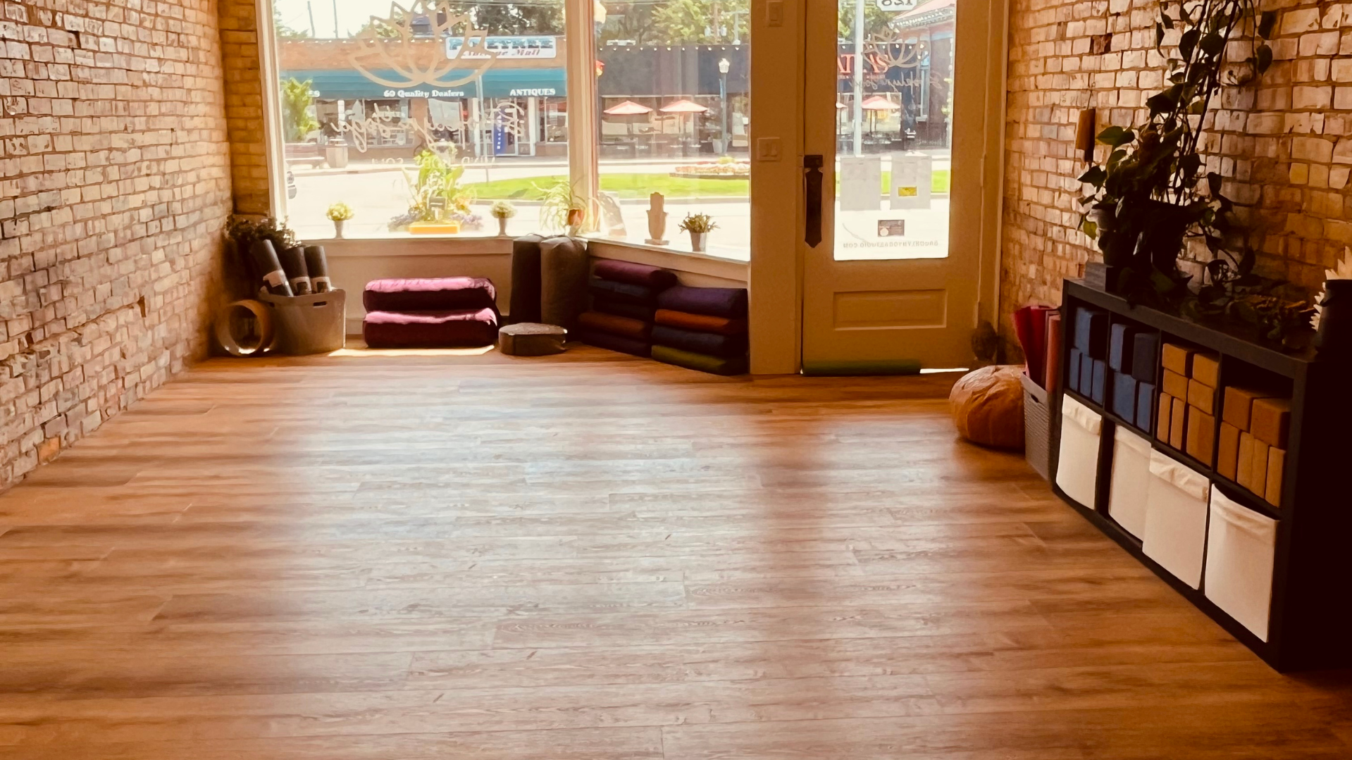 A welcoming yoga studio with wooden floor, exposed brick walls, large front window, and a door. Decor includes plants, yoga mats, yoga bolsters, baskets, and yoga props, with sunlight coming through the window and a view of Downtown Brooklyn, MI.