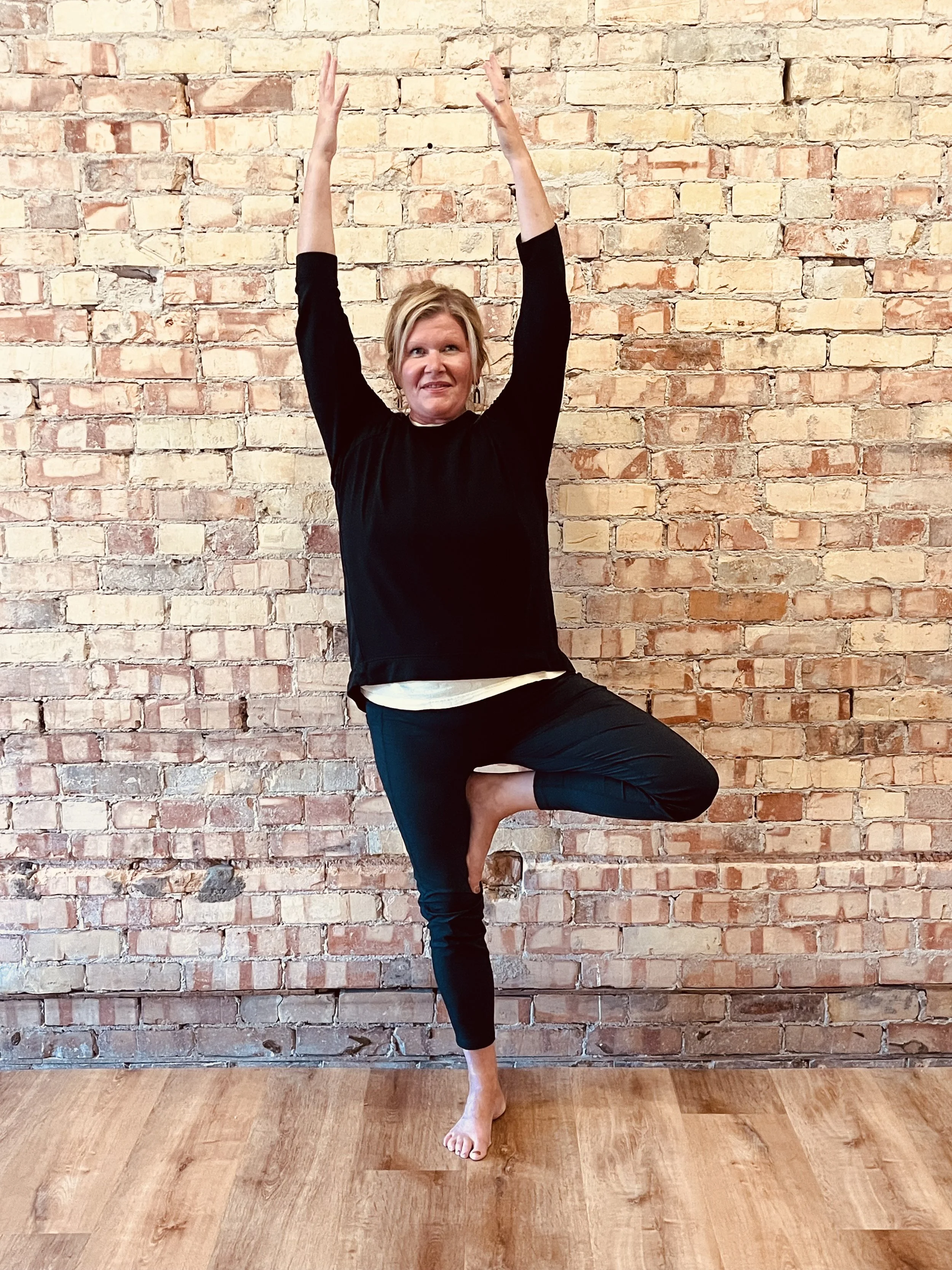 Woman standing on one leg in a yoga tree pose, with arms raised above her head and hands together, against a brick wall background.