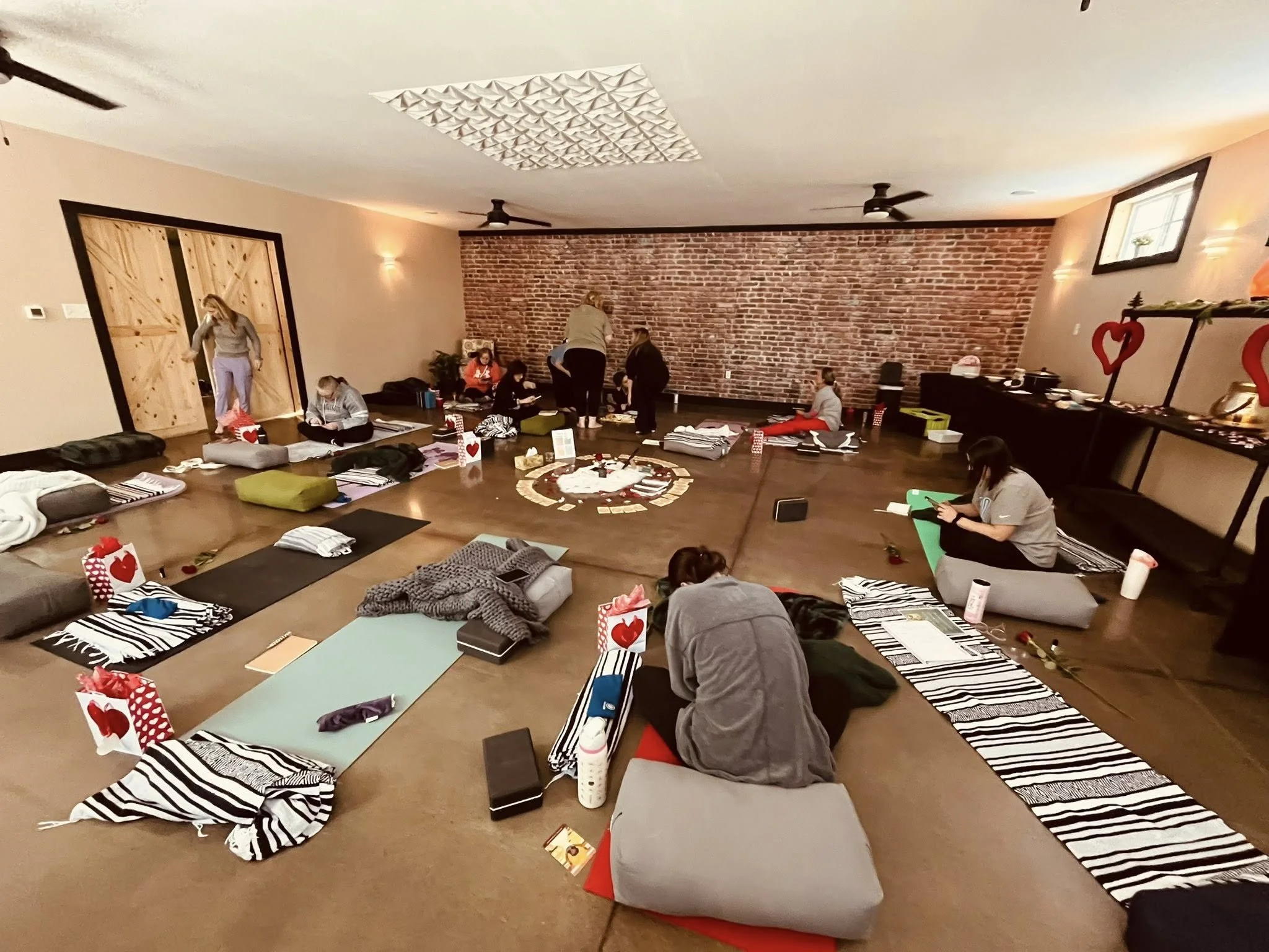 A group of yogis participating in a yoga retreat with a meditation class in a spacious studio with brick walls, yoga mats, pillows, and small gifts or decorations.