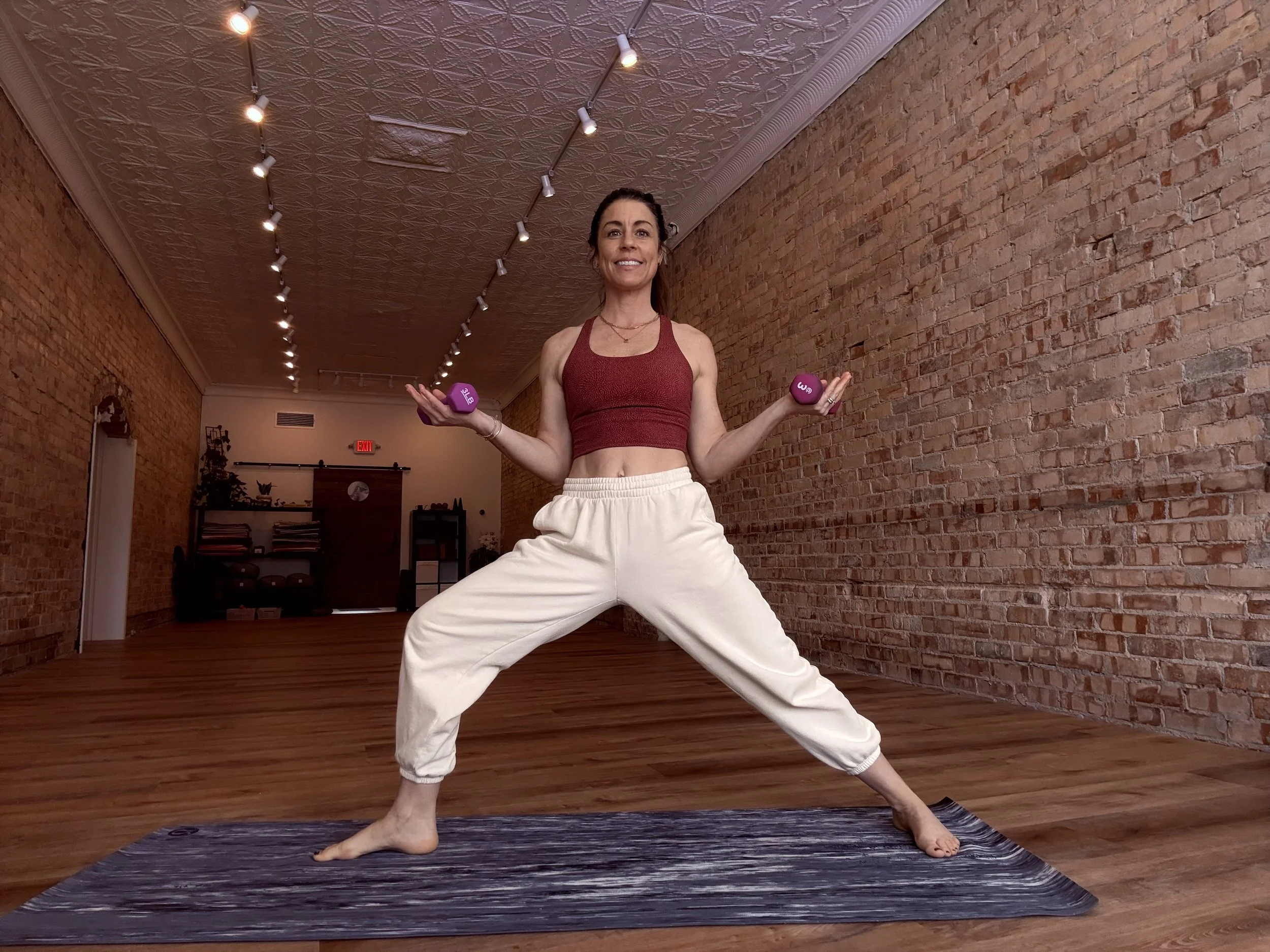 A woman practicing yoga in a spacious room with brick walls and wooden floors, standing on a yoga mat, holding small purple dumbbells, smiling, wearing a red sports bra and white sweatpants.