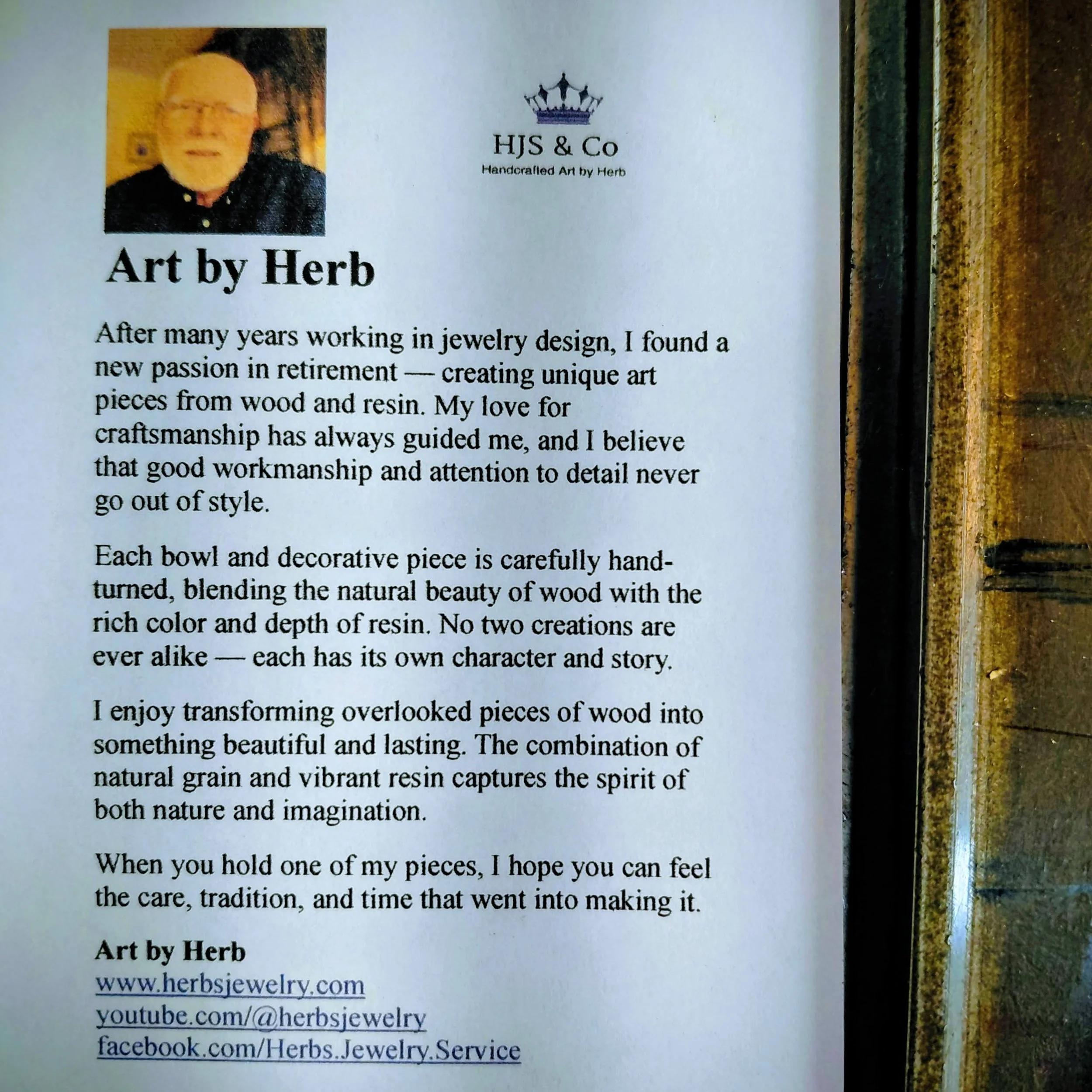 A close-up photograph of a printed artist's bio card for Herb, featuring his photo, logo, and description of his handcrafted wood and resin art, along with his website and social media links.