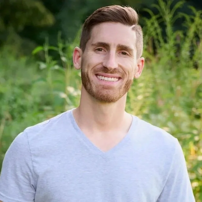 A portrait of a young man with brown hair, beard, and mustache, smiling, wearing a checkered shirt, against a plain white background.