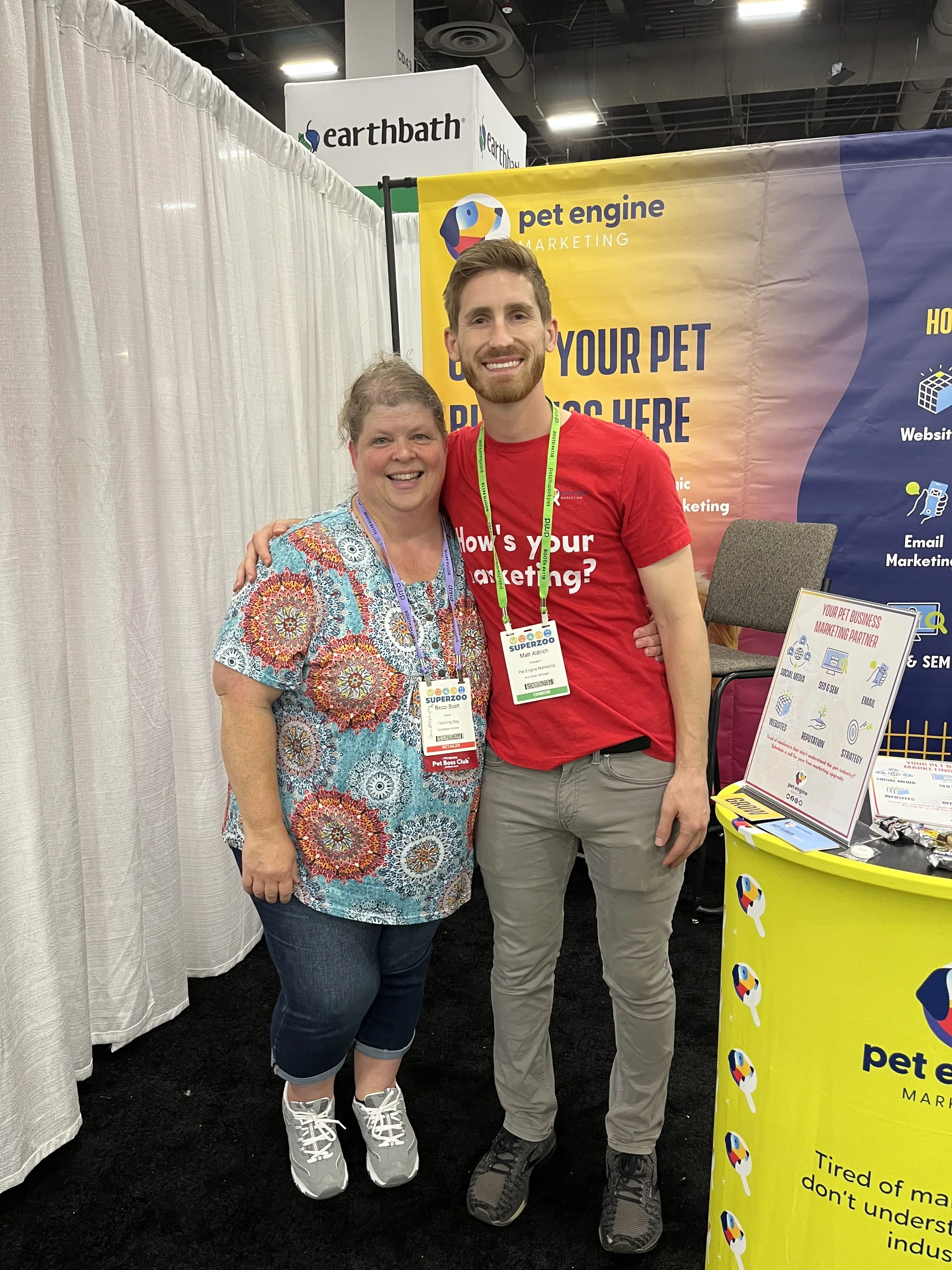 Two people smiling at a trade show booth for Pet Engine Marketing. The woman on the left is wearing a colorful floral shirt and jeans, and the man on the right is wearing a red T-shirt and gray pants. Behind them is a yellow and blue banner with the Pet Engine Marketing logo and information.